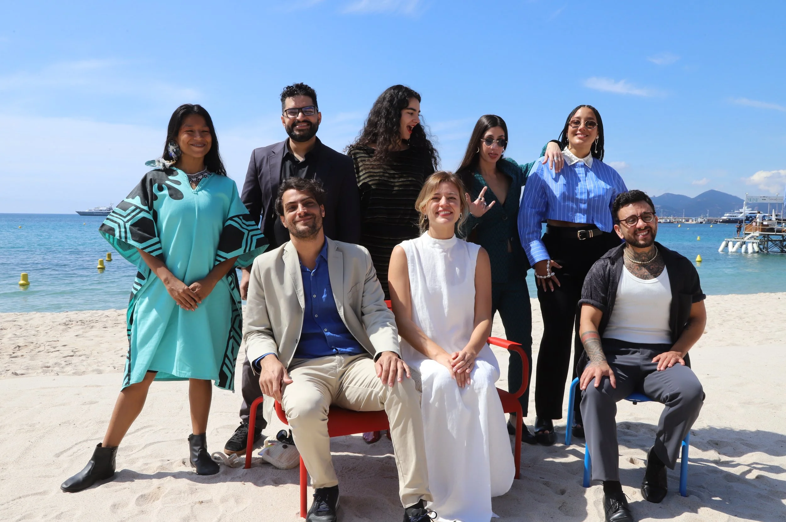 Group of eight diverse people at the beach, some sitting and some standing, with boats and mountains in the background on a sunny day.