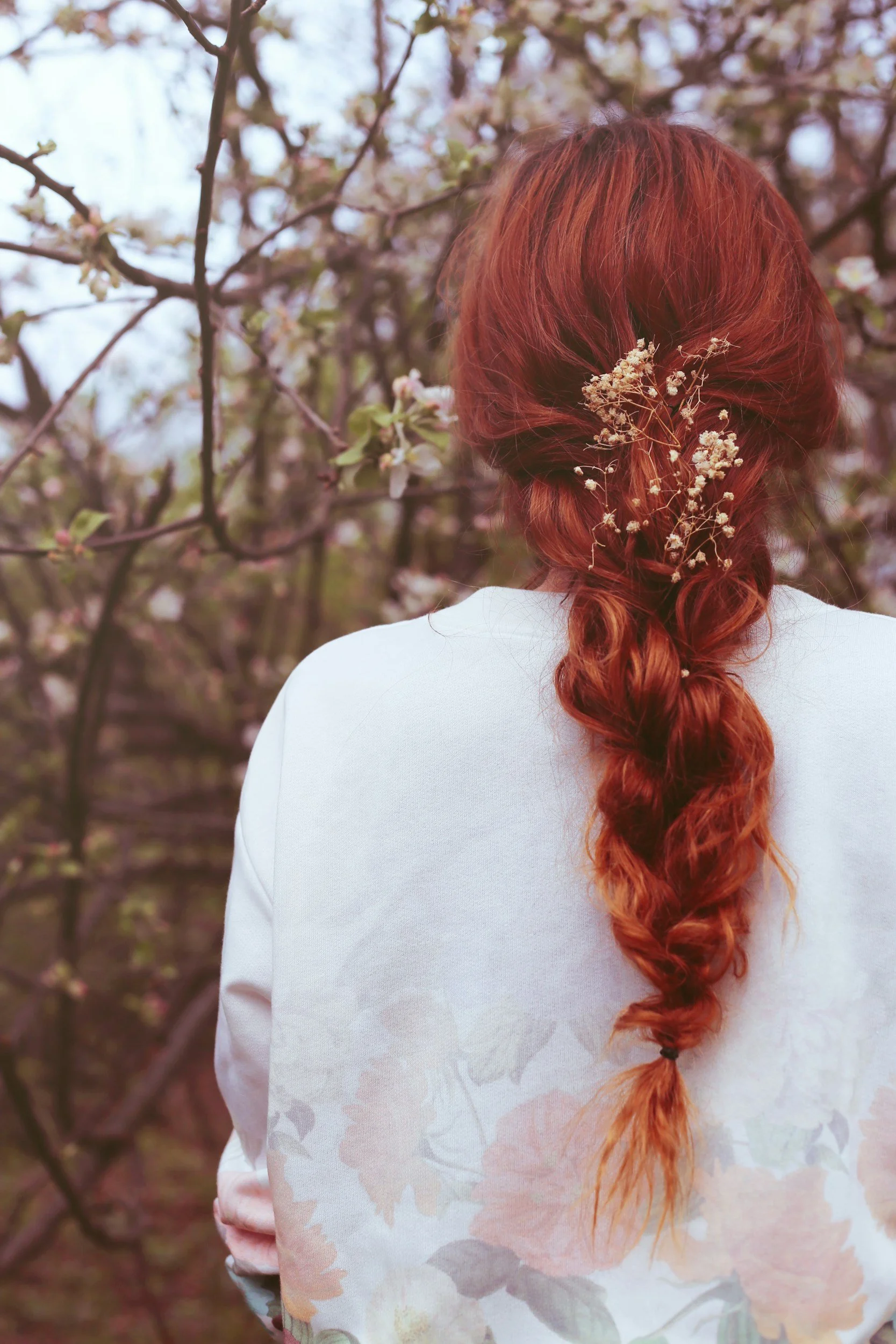 A woman with long, red, curly hair decorated with small white flowers, standing outdoors among flowering trees, with her back turned to the camera, wearing a white blazer.