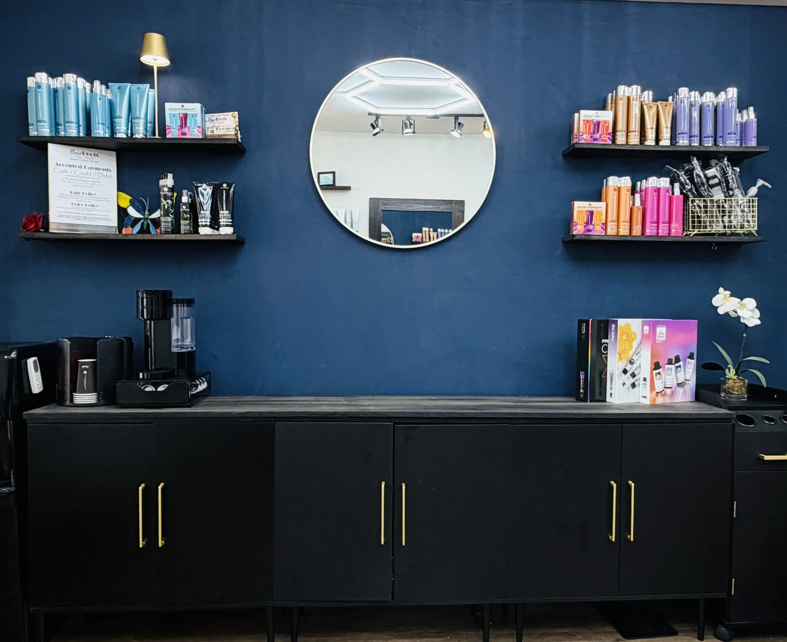 Bare Roots Hair Studio retail display with shelves of Keratherapy professional hair care products, a round mirror, black cabinetry, and a white orchid.