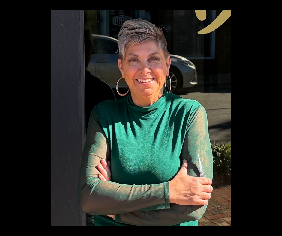 A smiling woman with short gray hair and hoop earrings stands outdoors in sunlight, wearing a green long-sleeved shirt with sheer sleeves, crossing her arms.