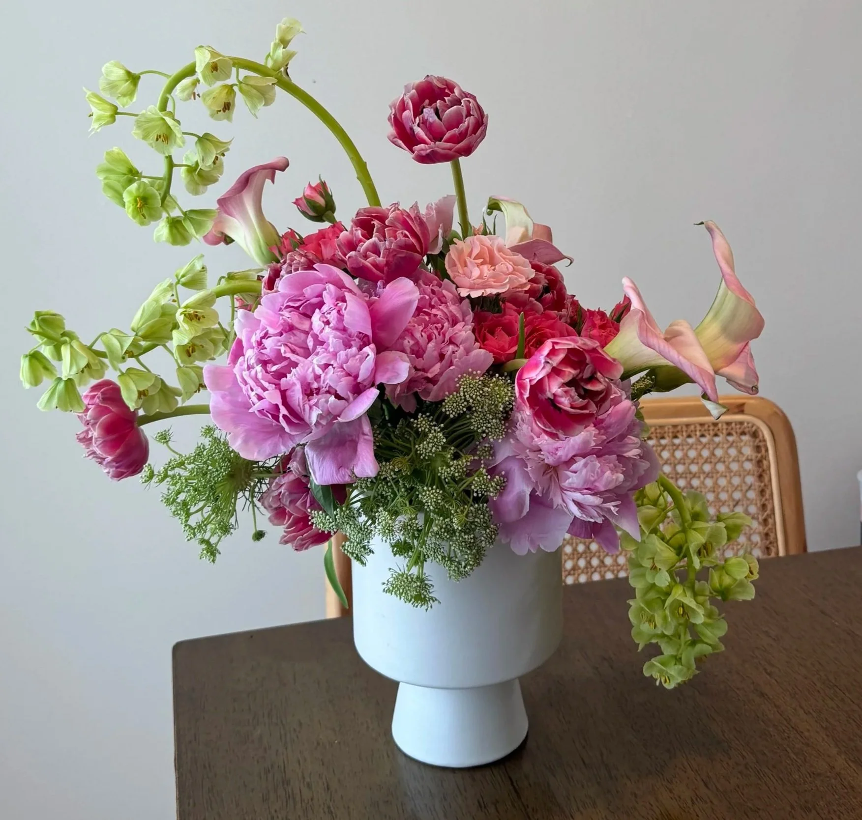 A bouquet of pink, purple, and green flowers in a white vase on a dark wooden table, with a rattan chair in the background.