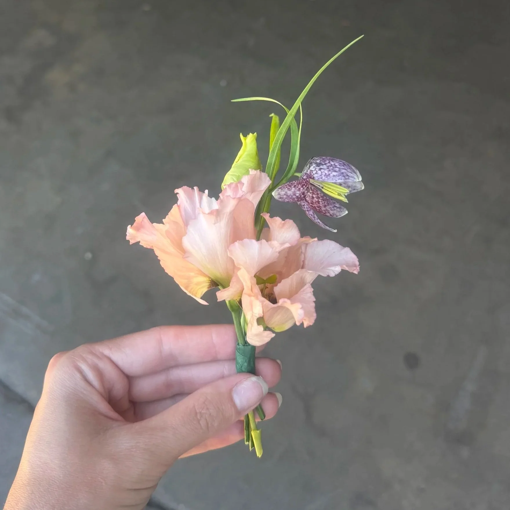 A hand holding a small bouquet of flowers against a gray background. The bouquet includes a peach-colored flower, a green bud, and a purple orchid.