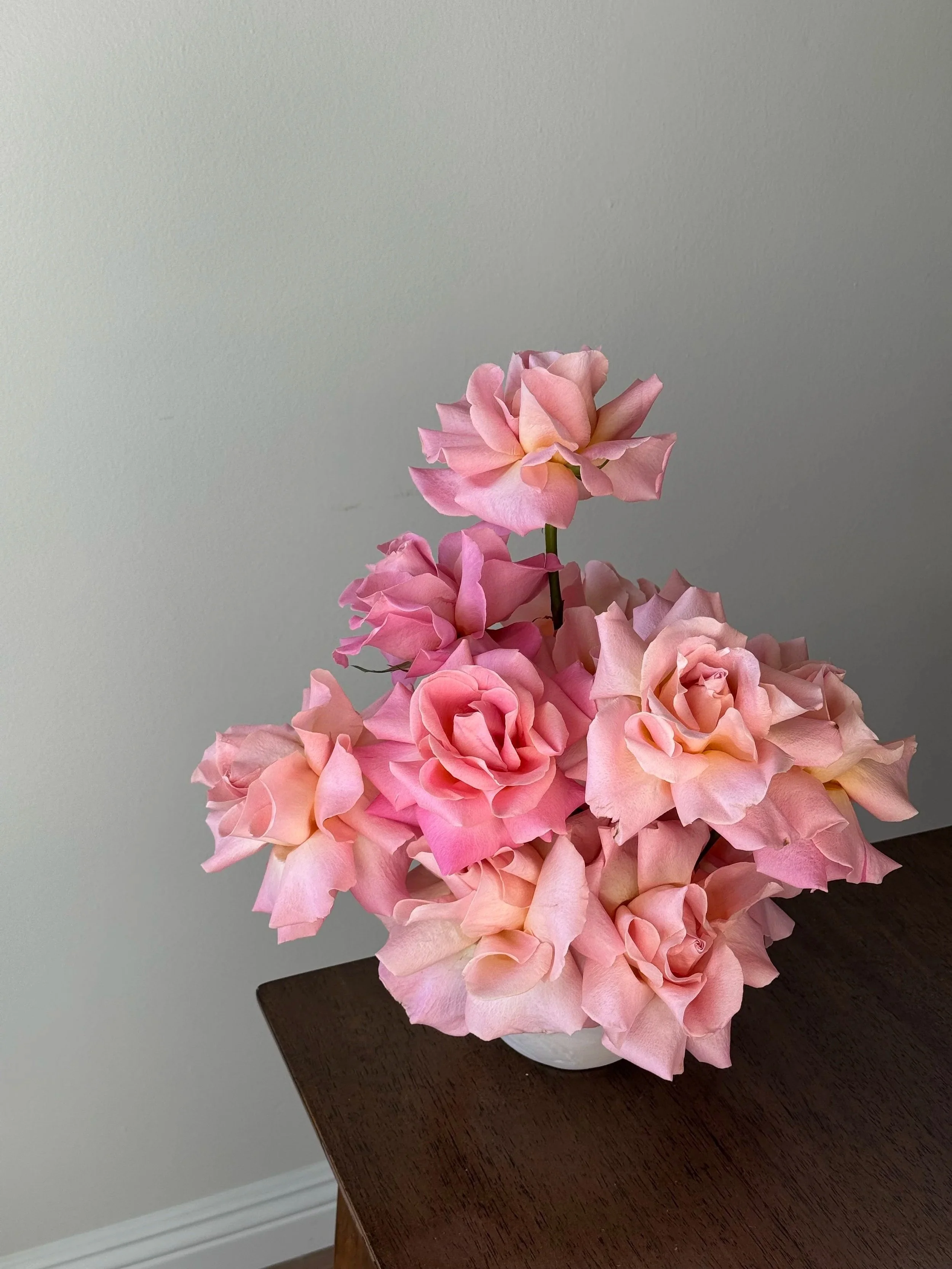 Pink and cream artificial roses in a white vase on a dark wooden table against a plain light-colored wall.