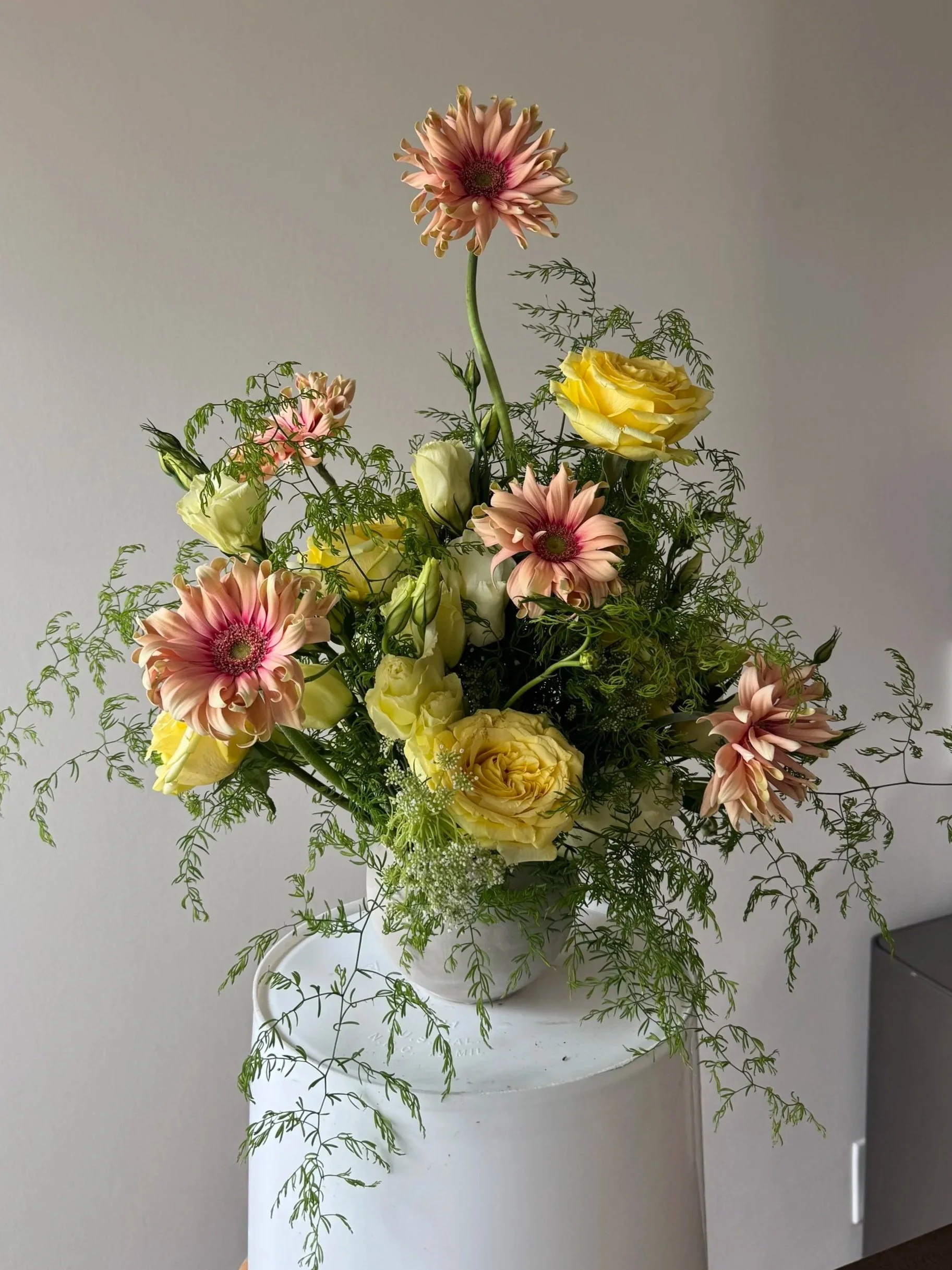 A tall floral arrangement in a white vase on a white surface, featuring pink gerbera daisies, yellow roses, and green foliage, against a plain gray background.