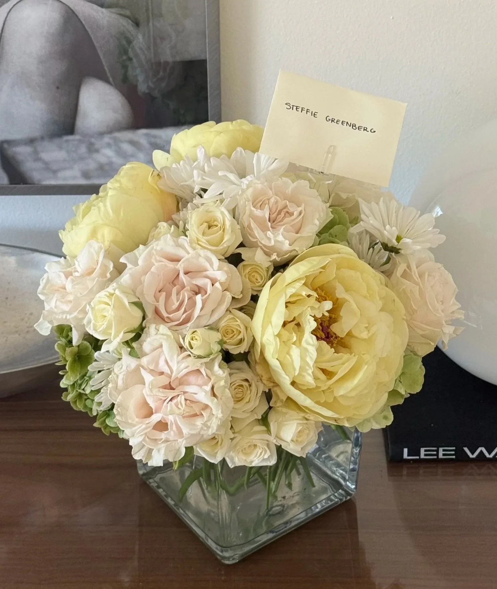 A bouquet of white, cream, and light yellow flowers in a clear glass vase on a dark wooden surface. Behind the flowers, there is a yellow card with handwritten text that reads "Steffie Greenberg." In the background, a reflective silver object and par