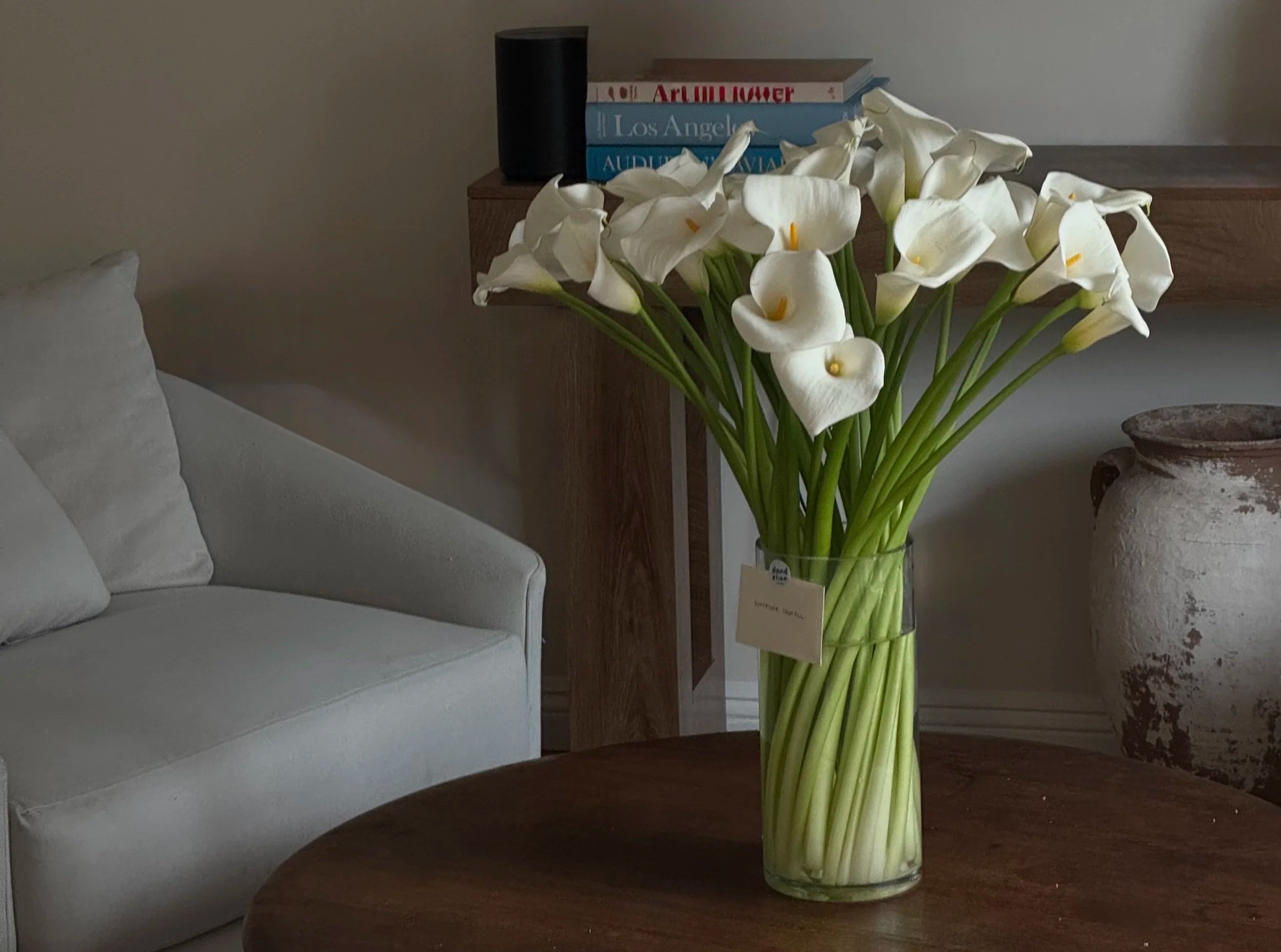 A glass vase of white calla lilies placed on a wooden table in a room with a white couch and a wooden side table with books and a black speaker.