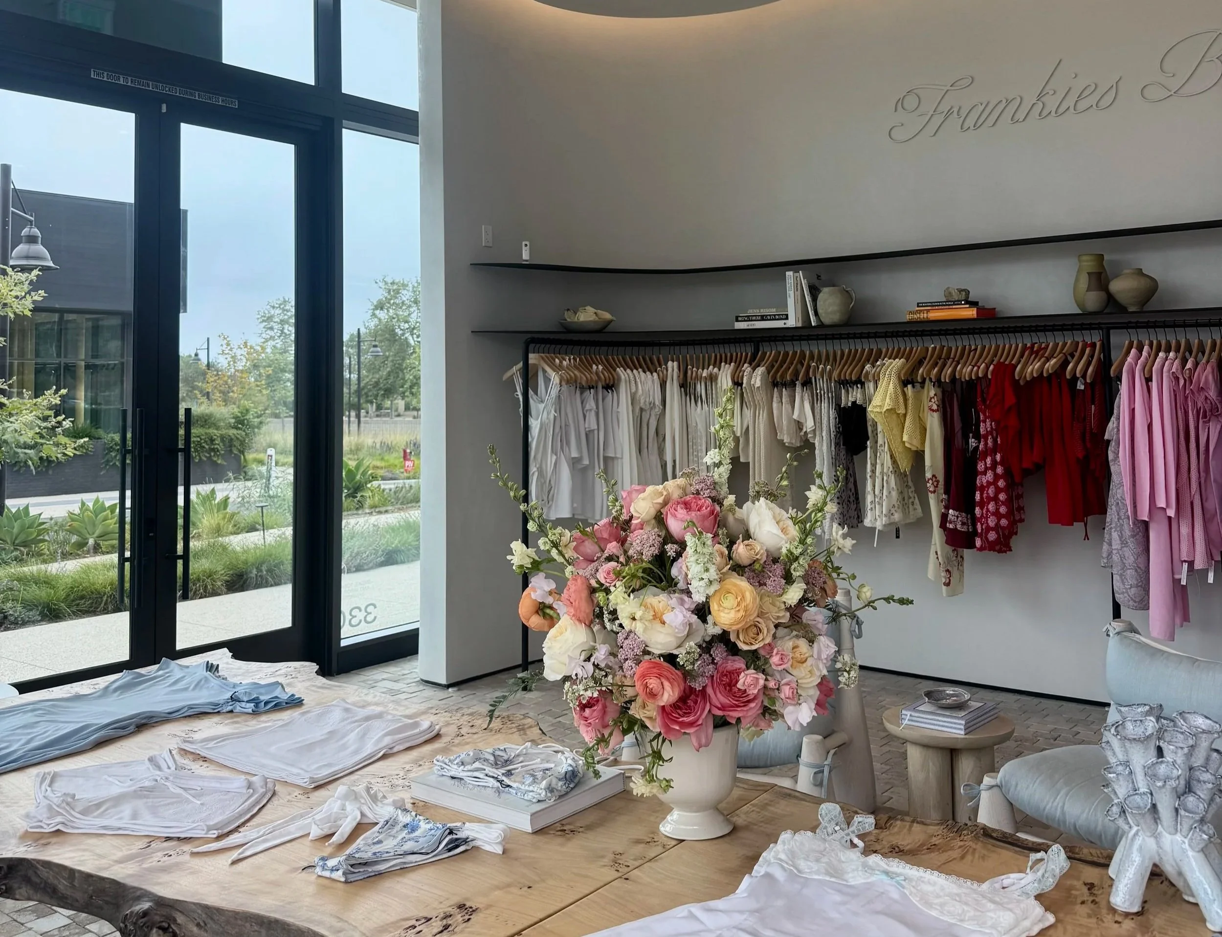 A retail store interior with a bouquet of pink and white flowers on a wooden table, various clothing items like T-shirts are laid out, and an apparel rack with dresses and tops. Large windows reveal outdoor greenery and a modern building.