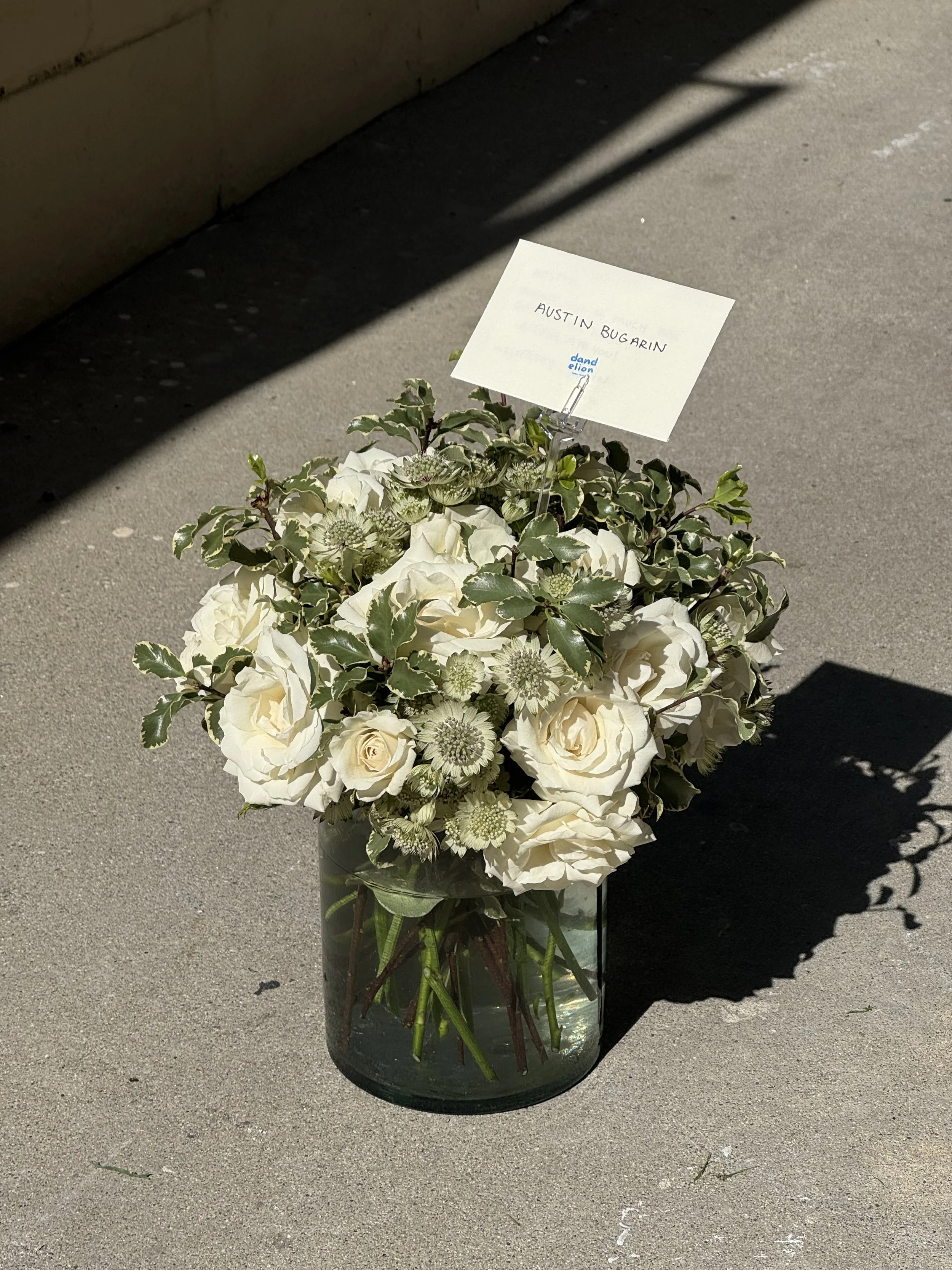 A bouquet of white flowers in a glass vase with a small sign that reads 'Austin Bugarin' and 'and elon,' placed on a concrete surface with a shadow cast across the image.