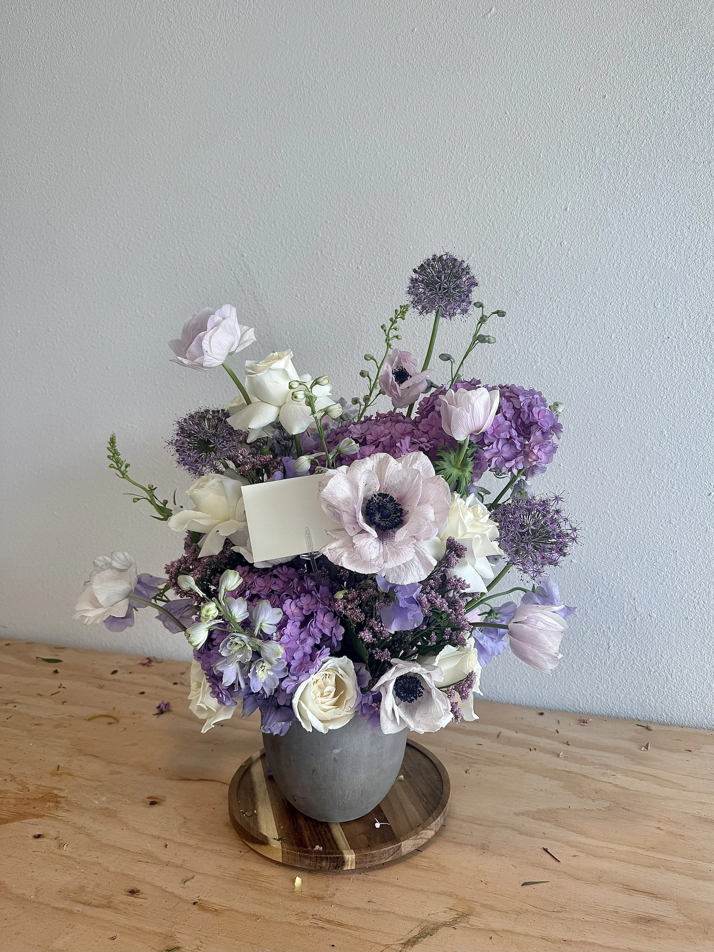 A bouquet of purple, white, and light pink flowers in a gray ceramic vase on a wooden table against a plain light-colored wall.