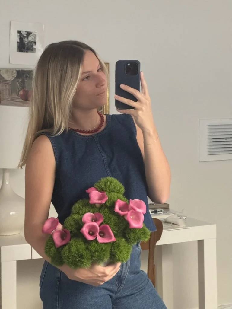 Woman with blonde hair taking a mirror selfie, holding a bouquet of pink calla lilies and green moss, inside a room with a white wall, framed photos, and a side table.