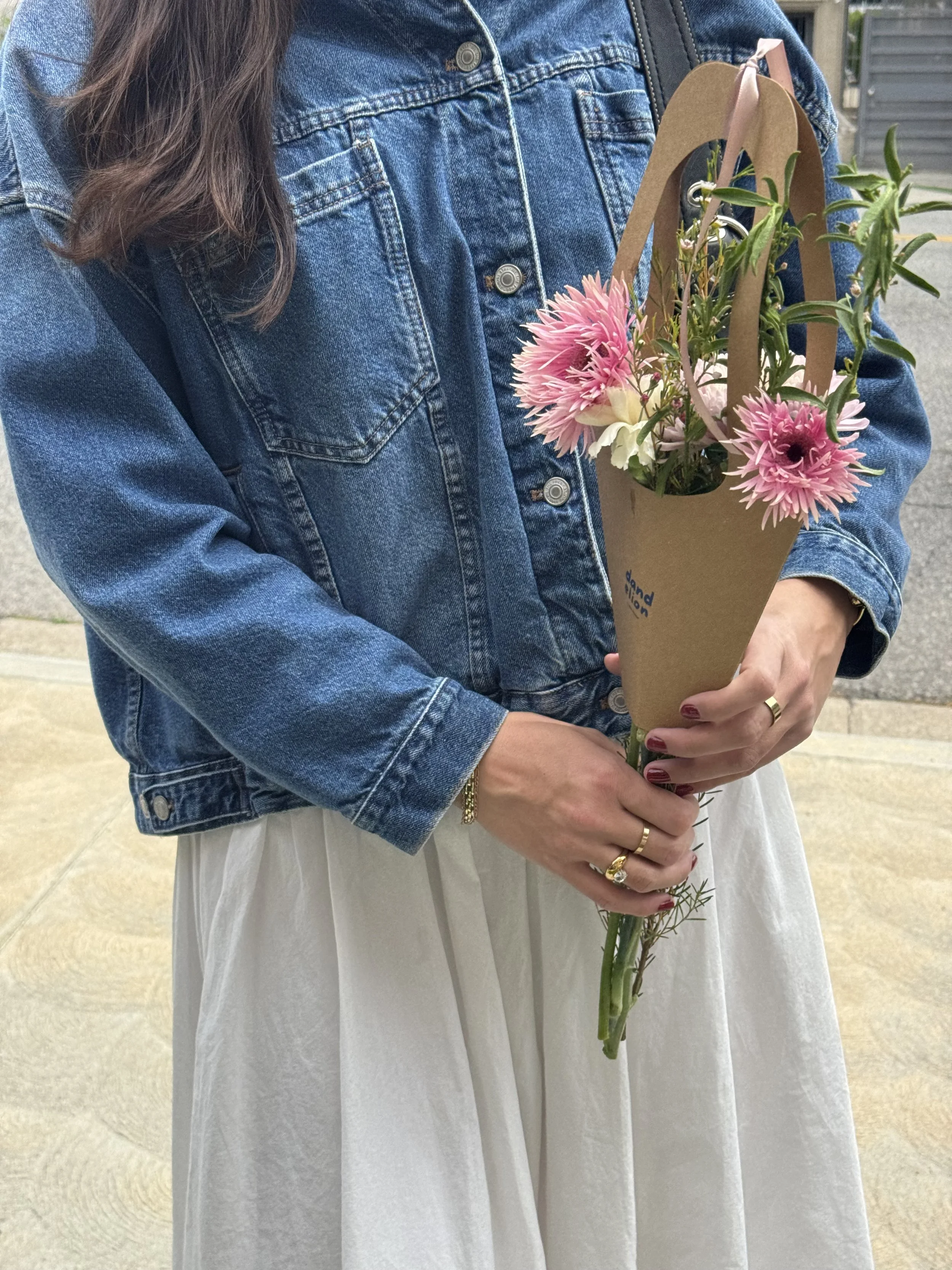 A person in a blue denim jacket and white dress holding a bouquet of pink and white flowers with green foliage, outdoors.