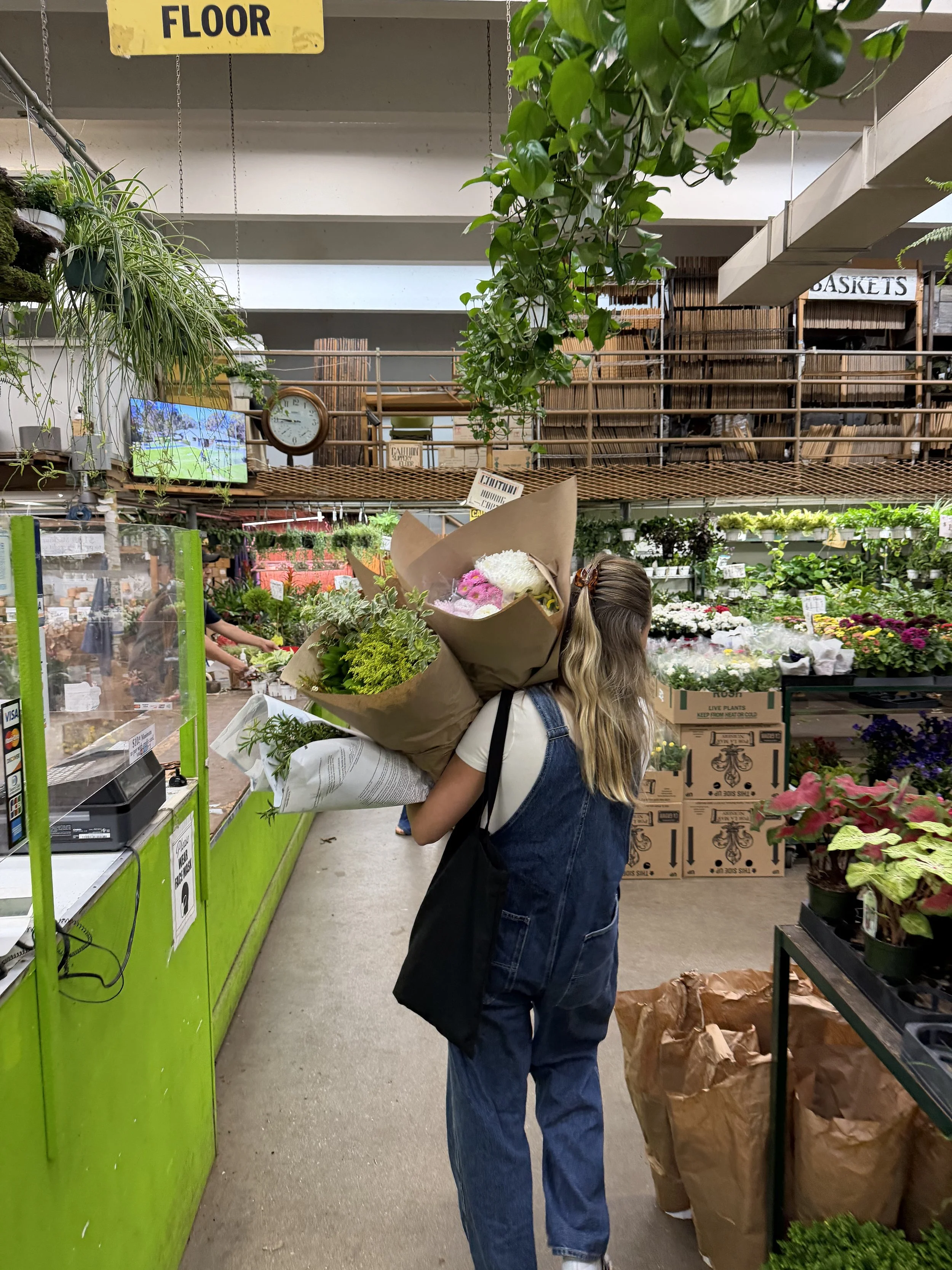 A woman holding a large bouquet of flowers inside a floral shop. She is wearing a white t-shirt and blue overalls, and her hair is tied back. There are various plants and flowers displayed on shelves around her, with a green counter and a checkout area in the foreground. A person in a black shirt can be seen in the background.