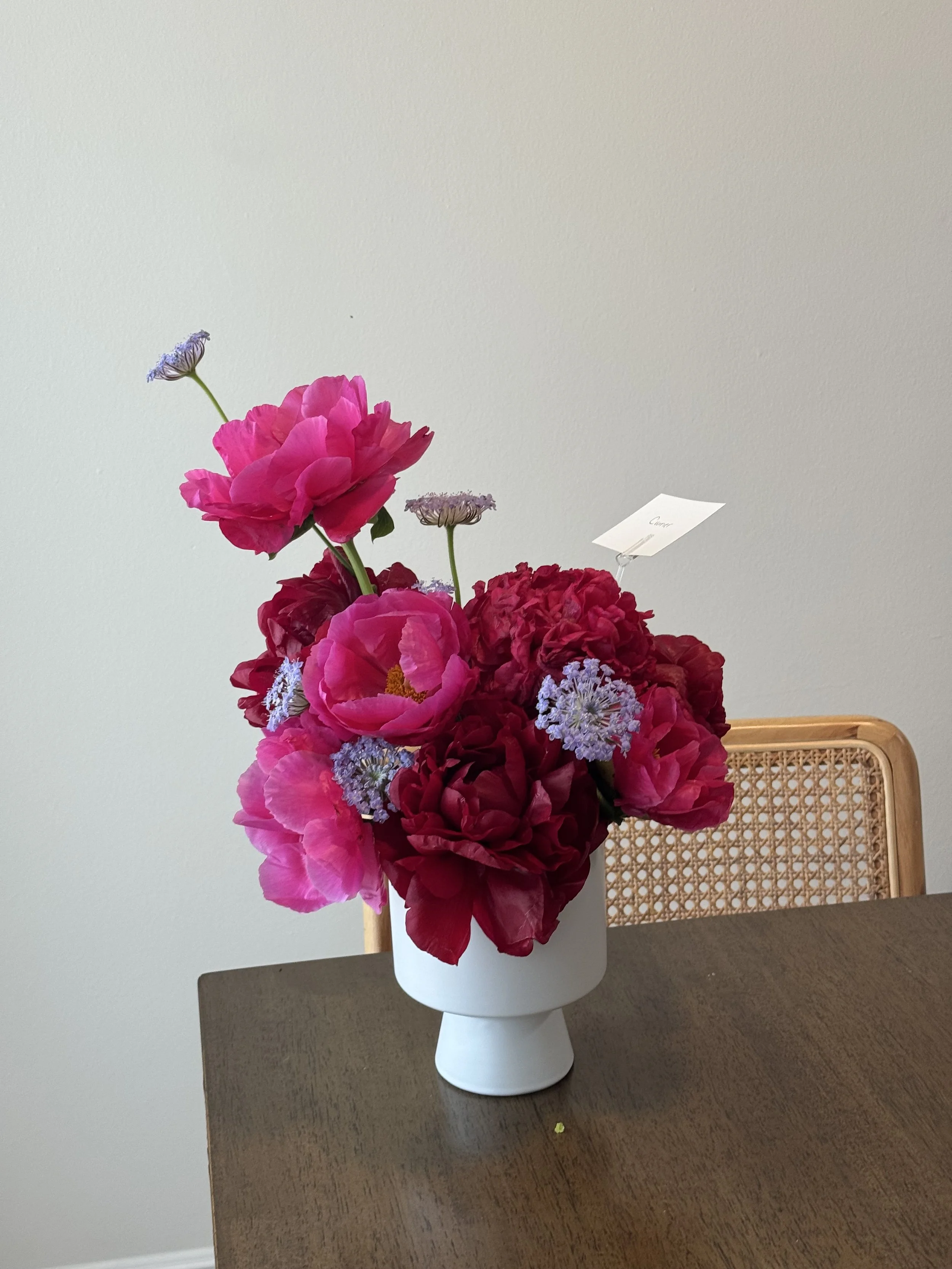 A bouquet of bright pink, deep red, and purple flowers in a white vase on a wooden table, with a chair partially visible behind it and a blank white wall in the background.