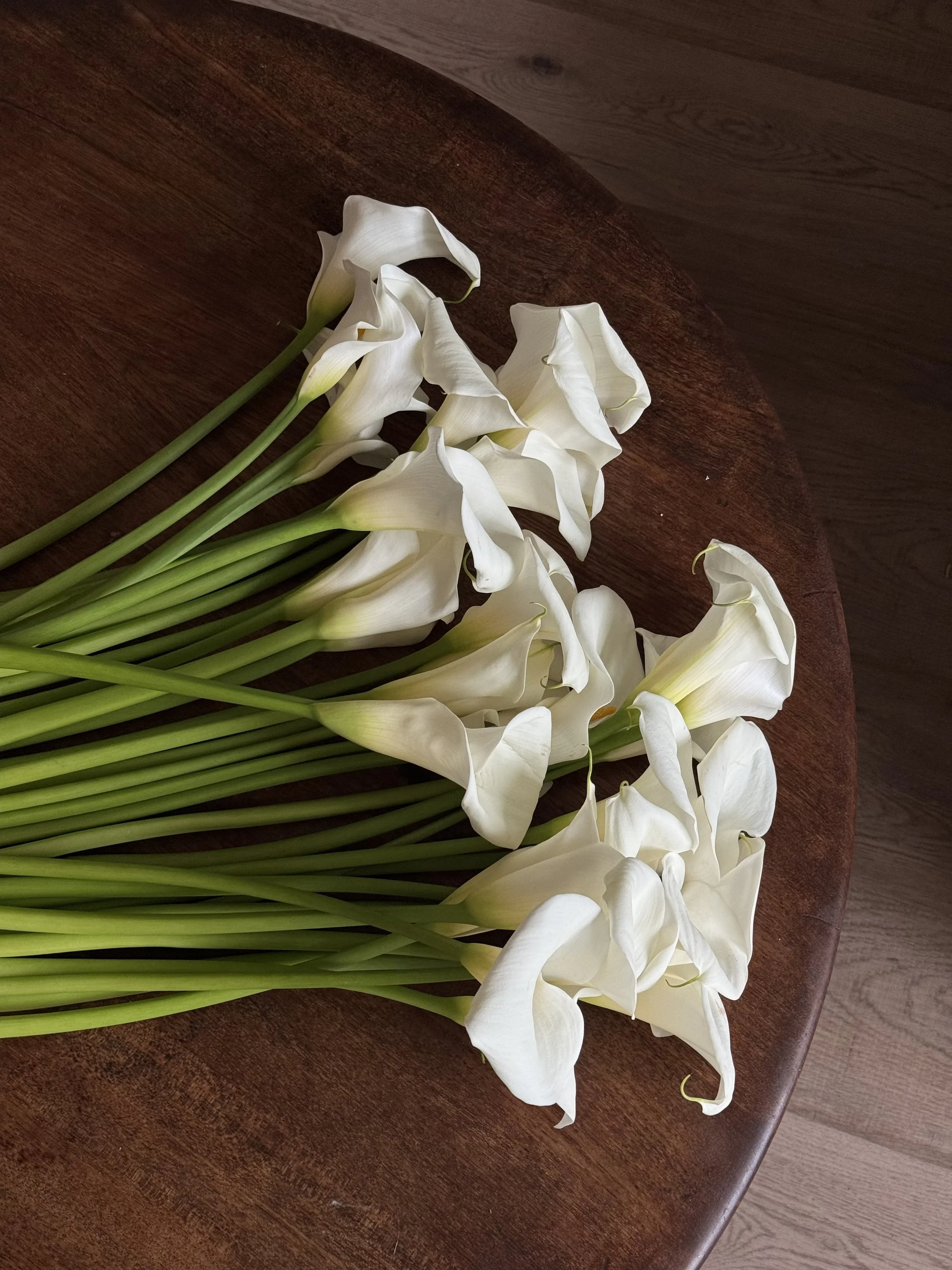 A bunch of white calla lilies with long green stems on a wooden surface.