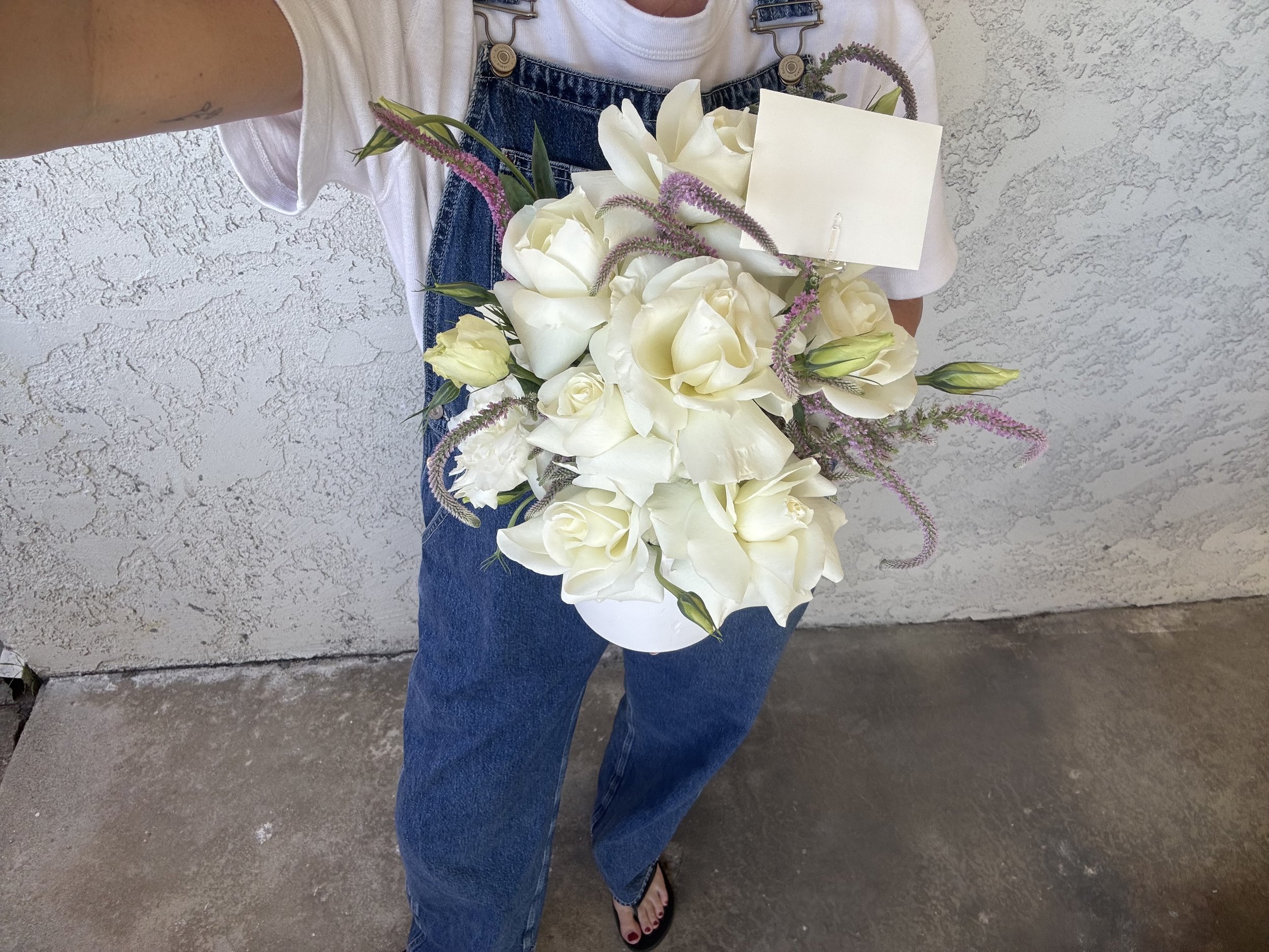 Person holding a bouquet of white roses with pink accents, standing against a textured wall, wearing a white t-shirt, blue overalls, and black flip-flops.