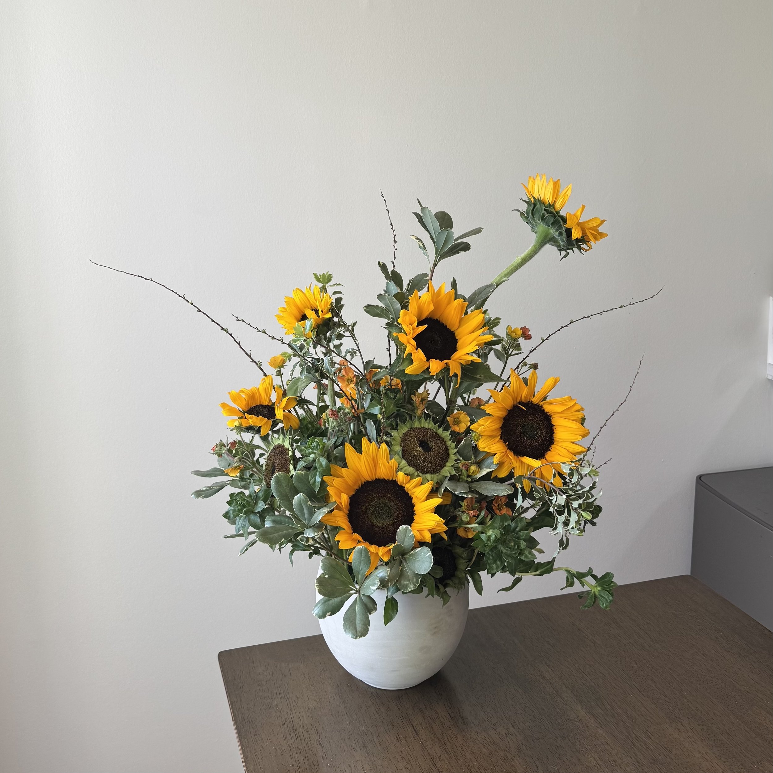 A bouquet of sunflowers and greenery in a white vase on a wooden table, against a neutral wall background.
