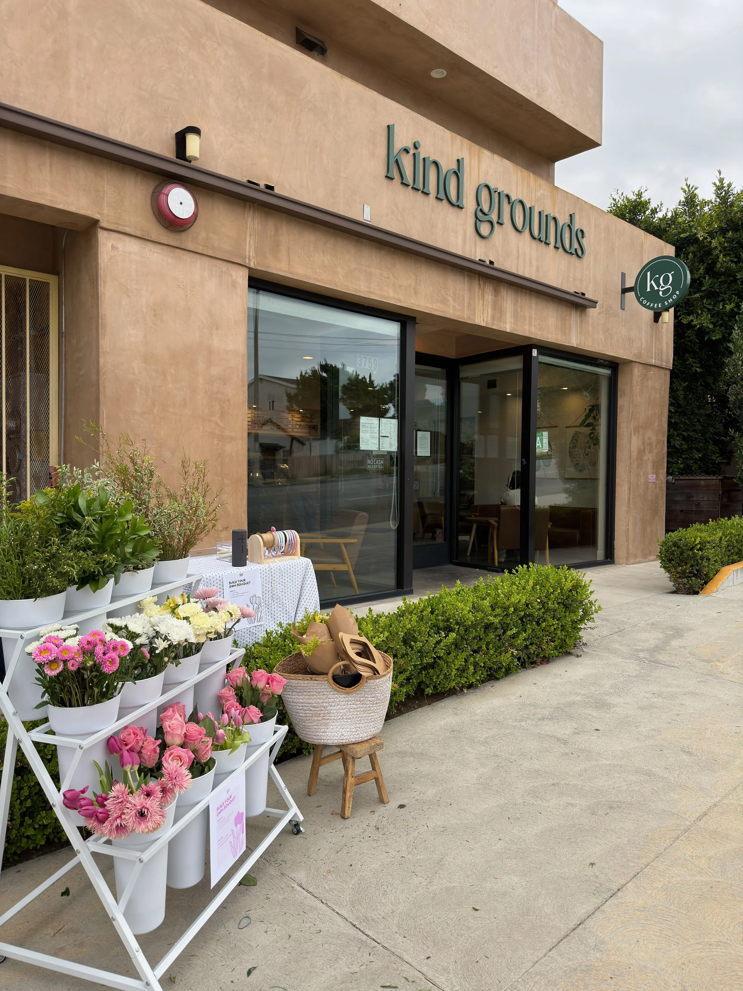 Flower stand outside a coffee shop named 'Kind Grounds'. The stand displays various pink and white flowers in white buckets, with additional flowers and a basket of beige bags on a small wooden stool nearby. The shop has large glass windows and a sig