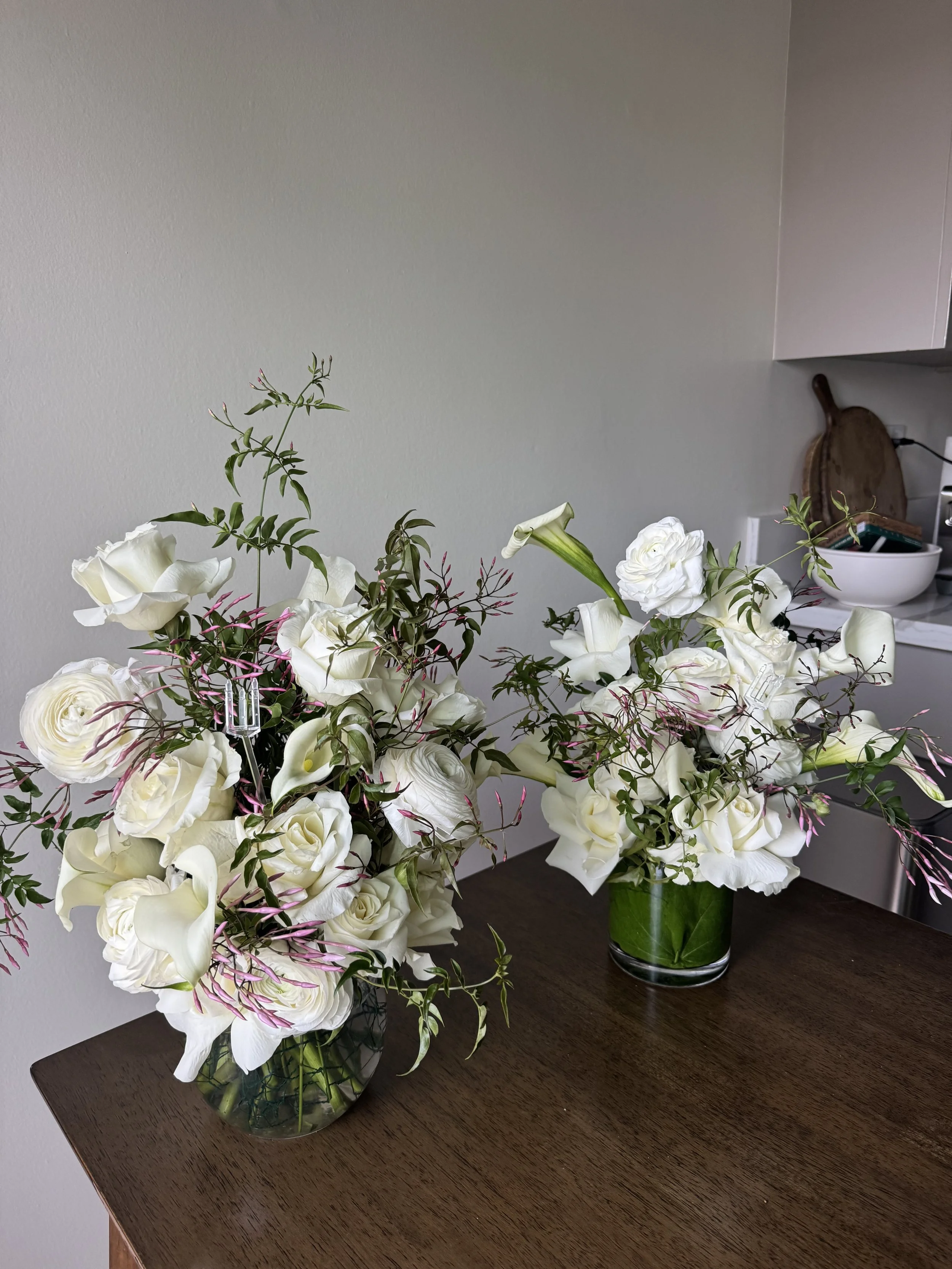 Two vases with white flower arrangements on a dark wooden table against a neutral wall, with a cutting board and white bowl in the background.