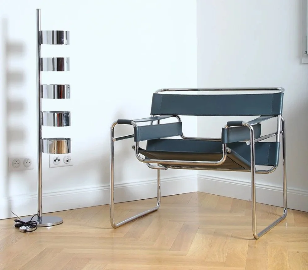Une chaise moderne en métal avec coussins bleus, placée dans un coin d'une pièce, à côté d'une lampe à pied en métal et d'une prise électrique sur le mur.