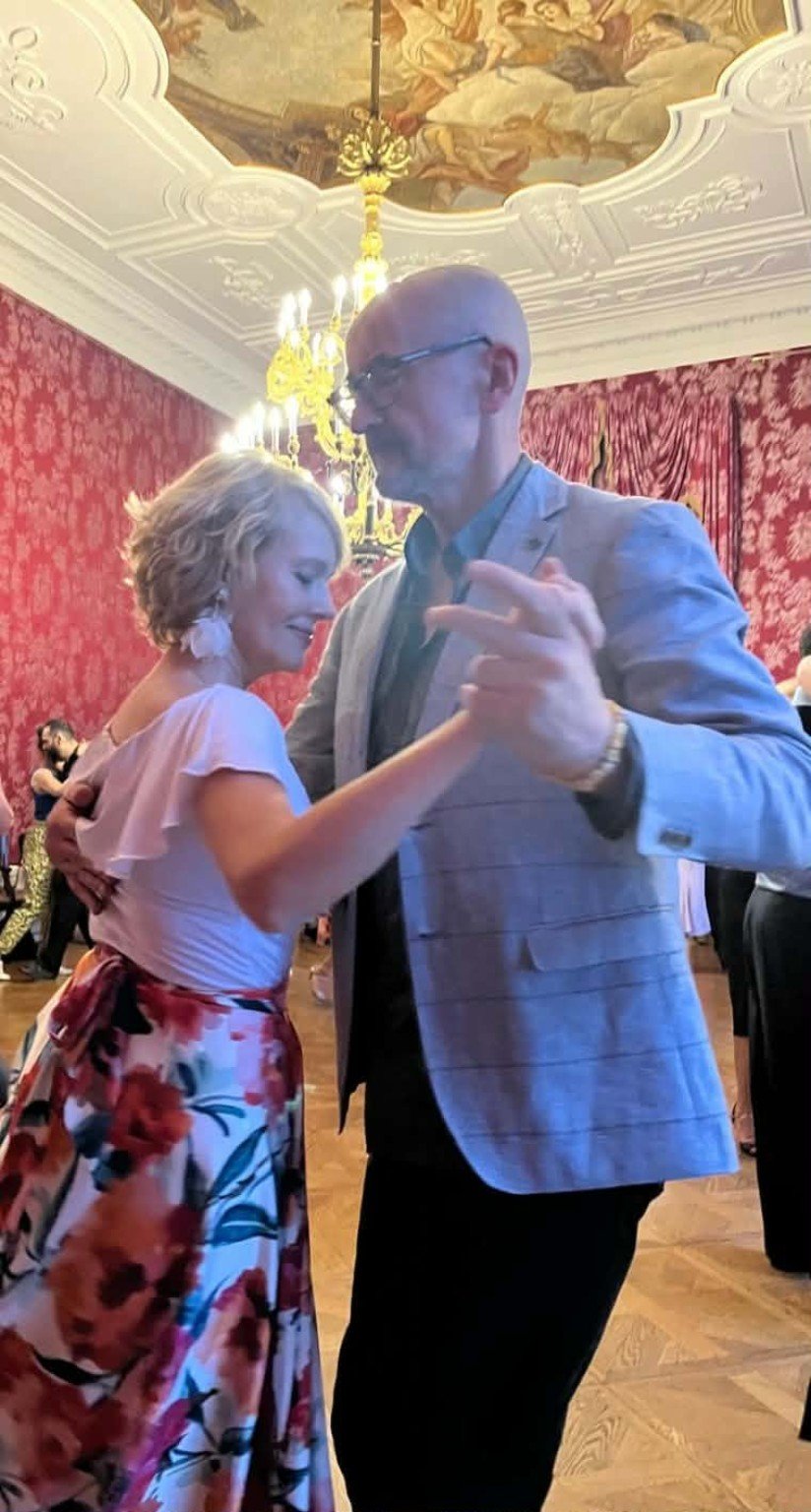 A man and a woman are dancing together in an elegant room with ornate white ceiling moldings, a chandelier, and red floral wallpaper. The woman has short blonde hair and is wearing a floral skirt and a light pink top. The man is bald, wearing glasses, and dressed in a light gray suit jacket with a dark shirt. They are sharing a dance and smiling softly.
