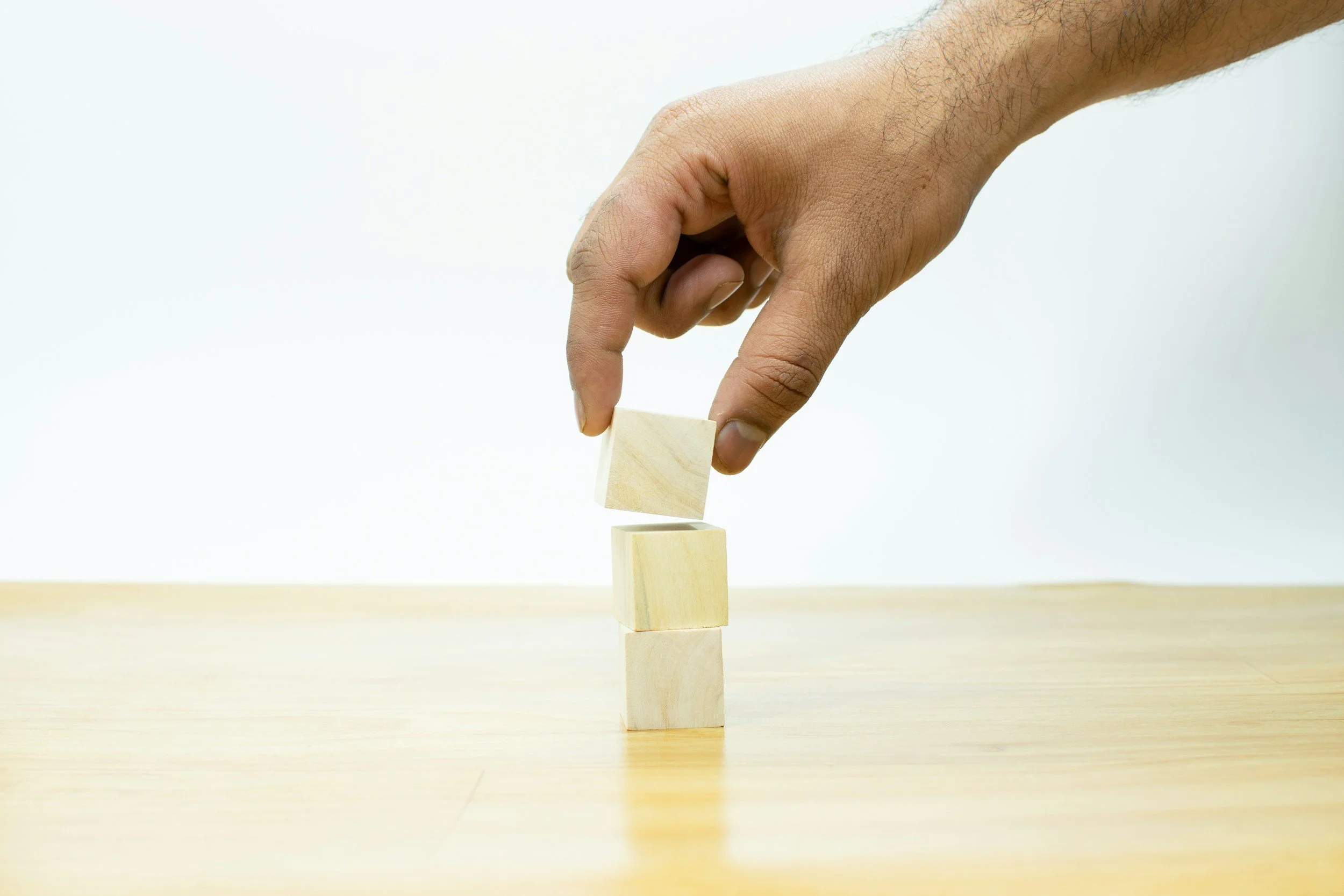 A hand stacking wooden blocks on a table, with three blocks already in place and one being added.