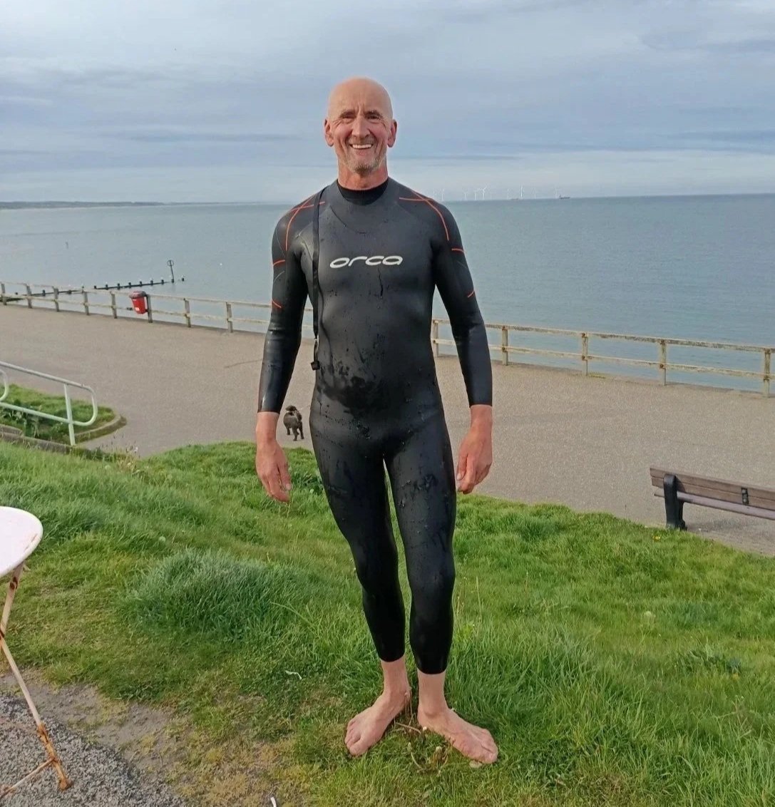 A man in a wetsuit standing on grass near a waterfront, smiling at the camera.