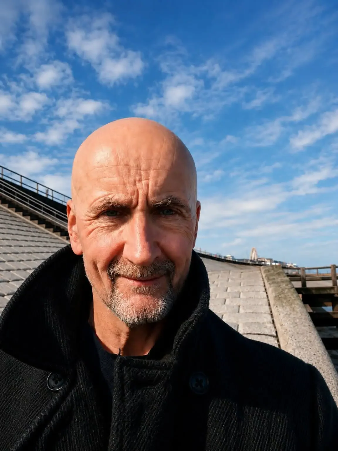 A close-up selfie of a smiling bald man with a gray beard, wearing a black jacket, taken outdoors on a sunny day with a clear blue sky and some clouds, along with a paved incline and a railing in the background.