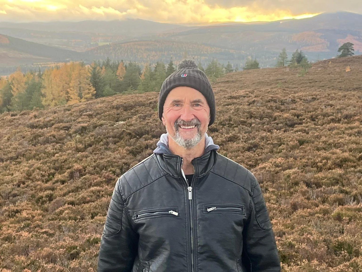 A smiling man wearing a black leather jacket and a black knit cap stands outdoors in a hilly landscape with trees and rolling hills in the background at sunset.