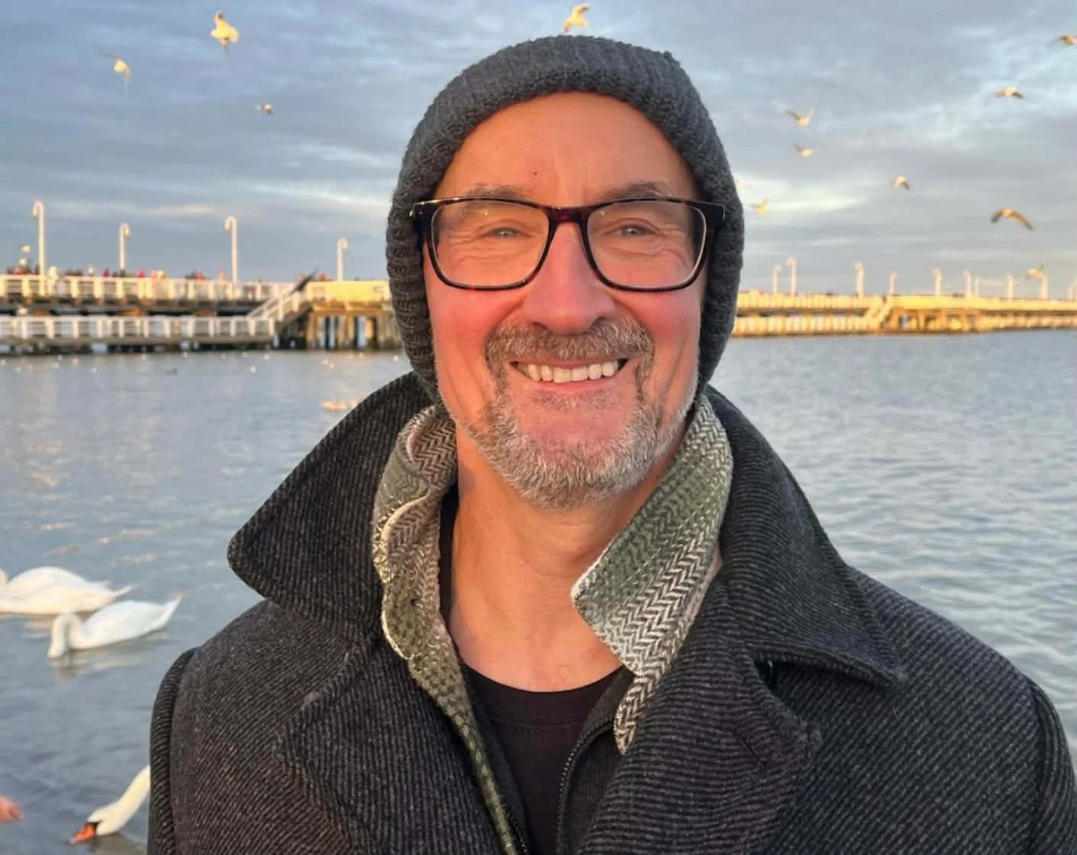 A smiling man with glasses, a beard, and a gray beanie, standing outdoors near a body of water with swans and a dock in the background.