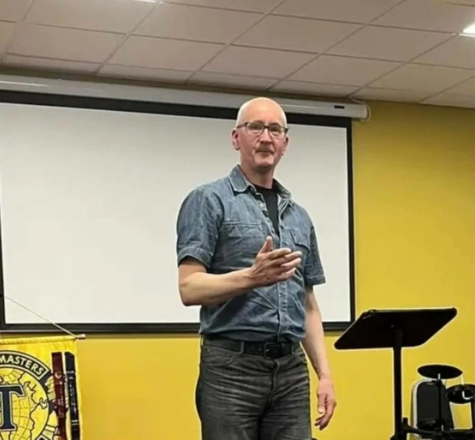 A man wearing glasses, a denim shirt, and dark pants speaking in front of a yellow wall with a projector screen behind him. There is a microphone stand and a music stand in front of him, and a banner with the Lions Club emblem on the left side of the image.