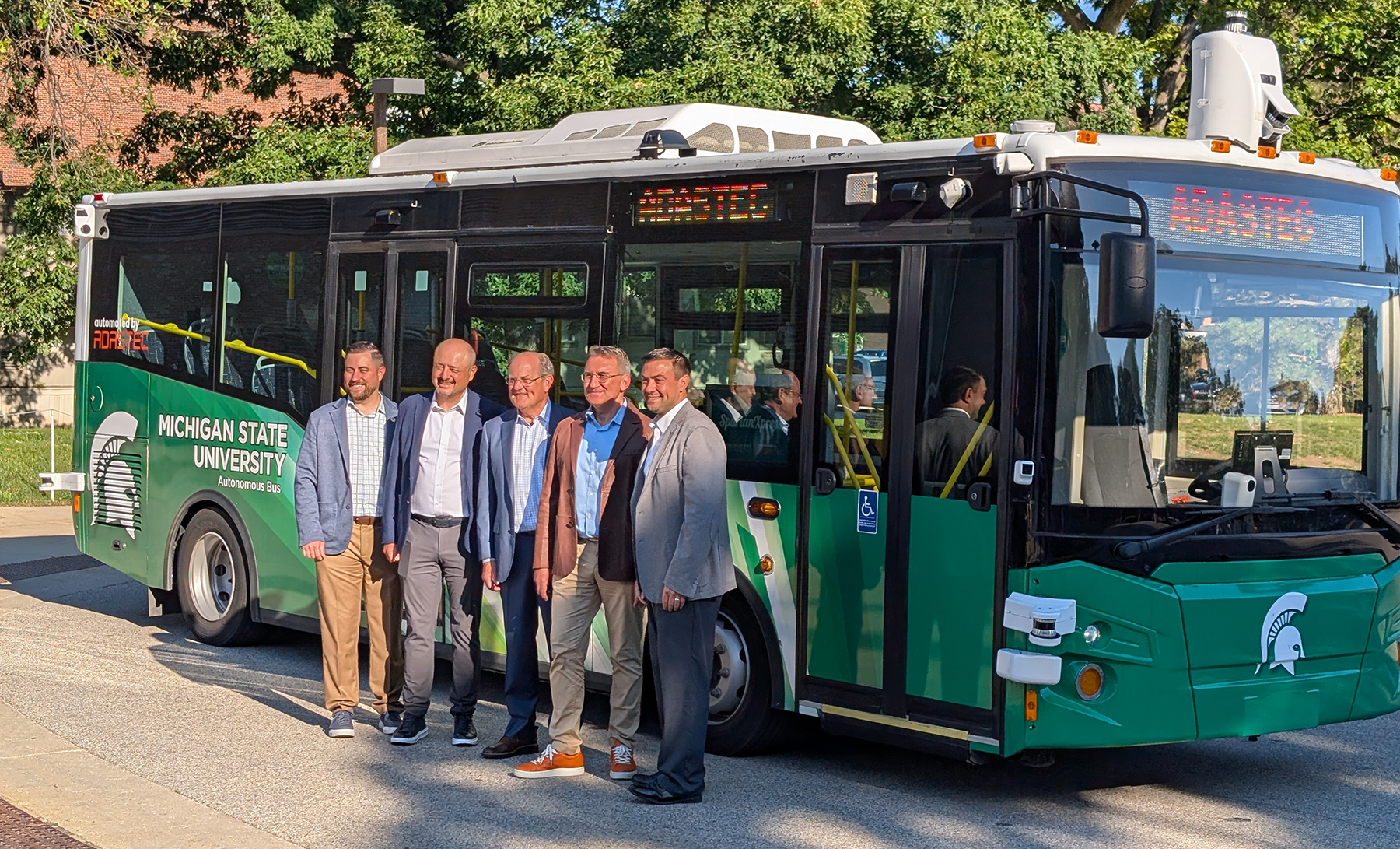 A group of five white men stand smiling in front of a green bus showing the MIchigan State University logo