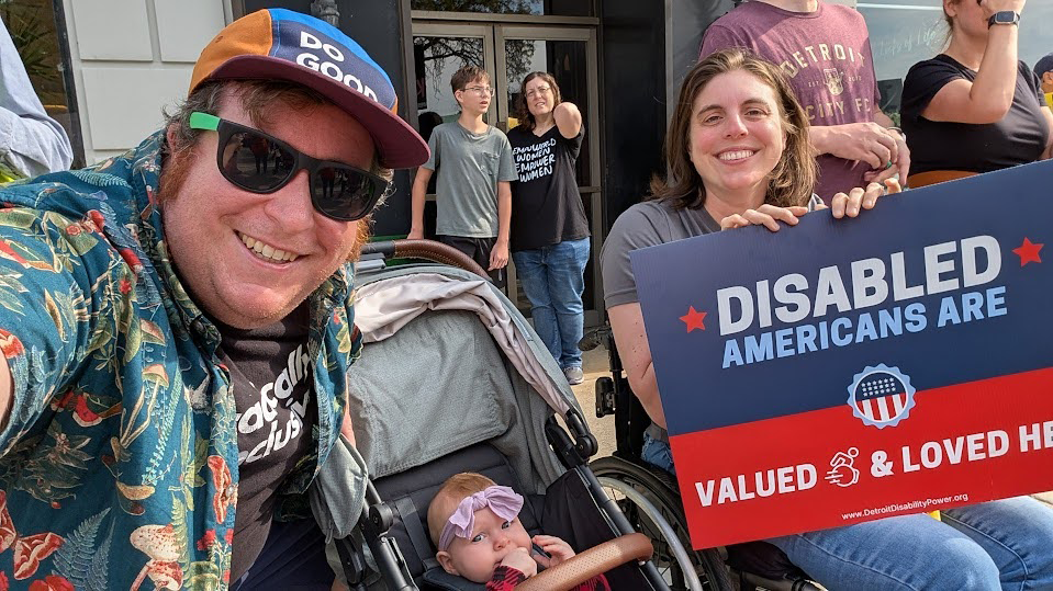 Group of people at a rally holding a sign that reads 'Disabled Americans are valued and loved here.' A man in sunglasses and a cap takes a selfie, a woman in a wheelchair holds the sign, and a baby in a stroller is present.