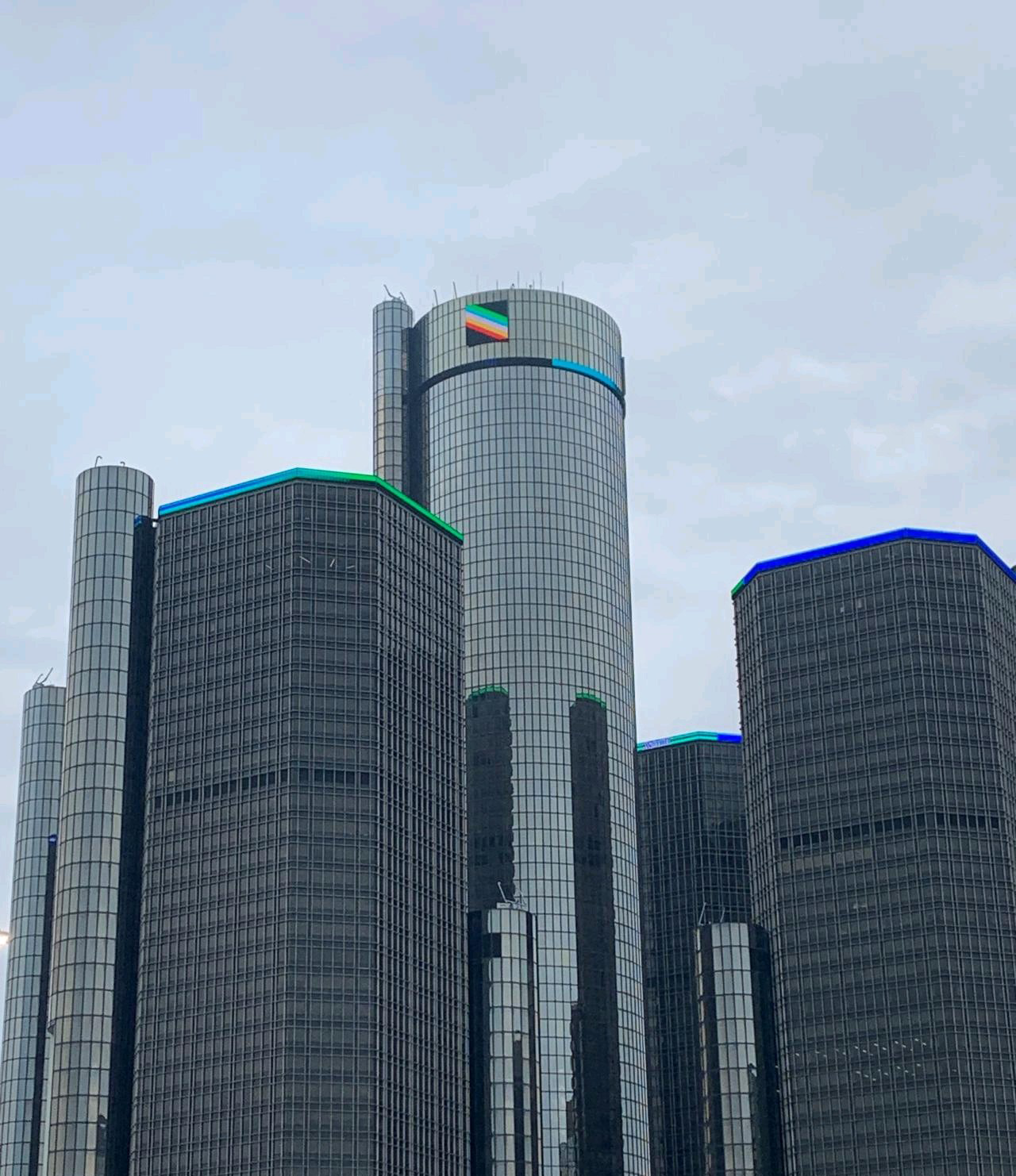 Tall, modern skyscrapers in the Detroit skyline with glass exteriors, showing a digital banner of the disability pride month flag at the top.