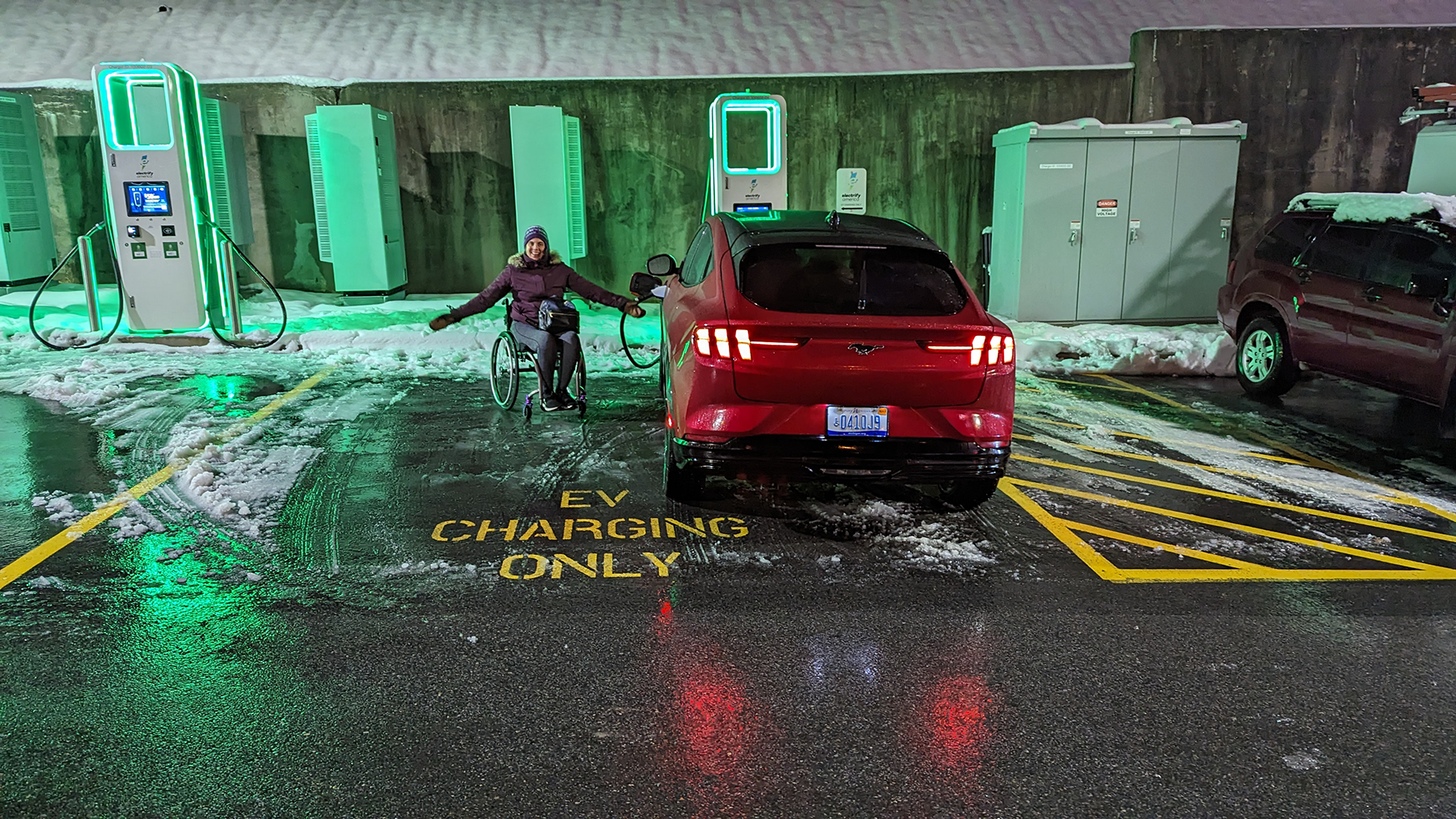 A woman in a wheelchair is next to her electric vehicle, charging it with her arms spread wide, gesturing to how wide the "EV charging only" parking space is, demonstrating ample accessibility