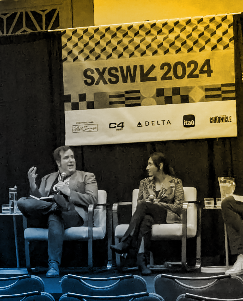 Alan Hejl speaking on a panel at SXSW 2024. He is an adult white cis male seated next to a woman in white chairs, in a panel setting.