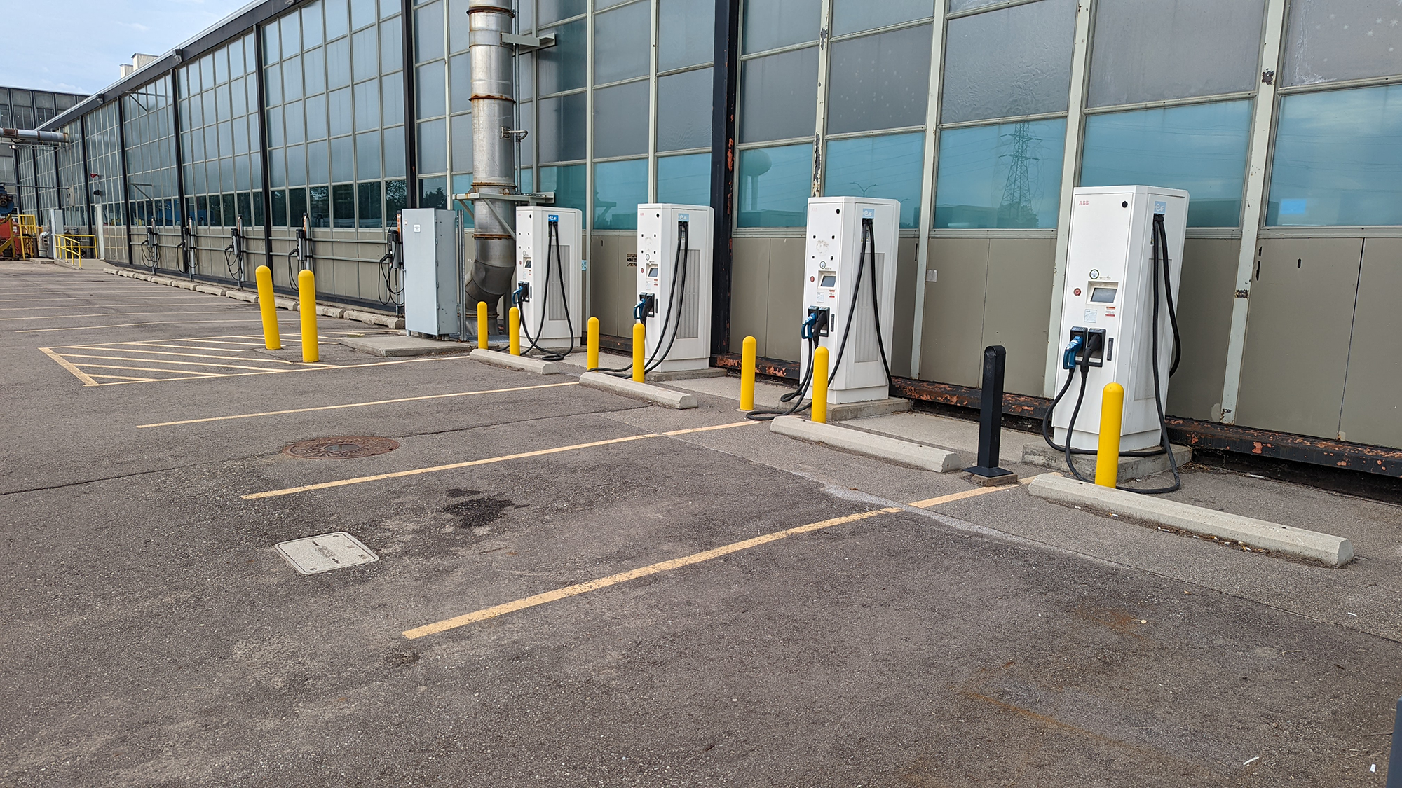 An outdoor parking lot with five electric vehicle charging stations along a building with large glass windows. The stations are white with digital screens and black charging cables. Yellow bollards protect each station, and the parking spaces are out
