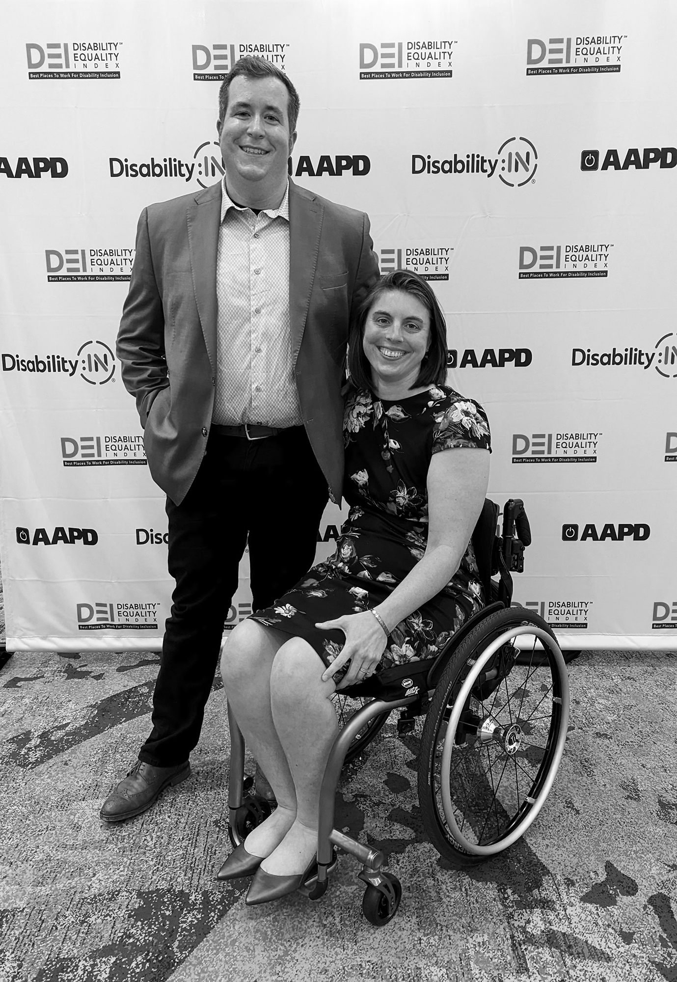 Alan Hejl standing next to his wife, a wheelchair user, in front of a 'step & repeat' at an event.