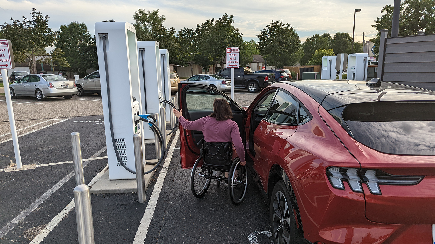 A woman in a wheelchair charging her red car at an electric vehicle charging station in a parking lot.