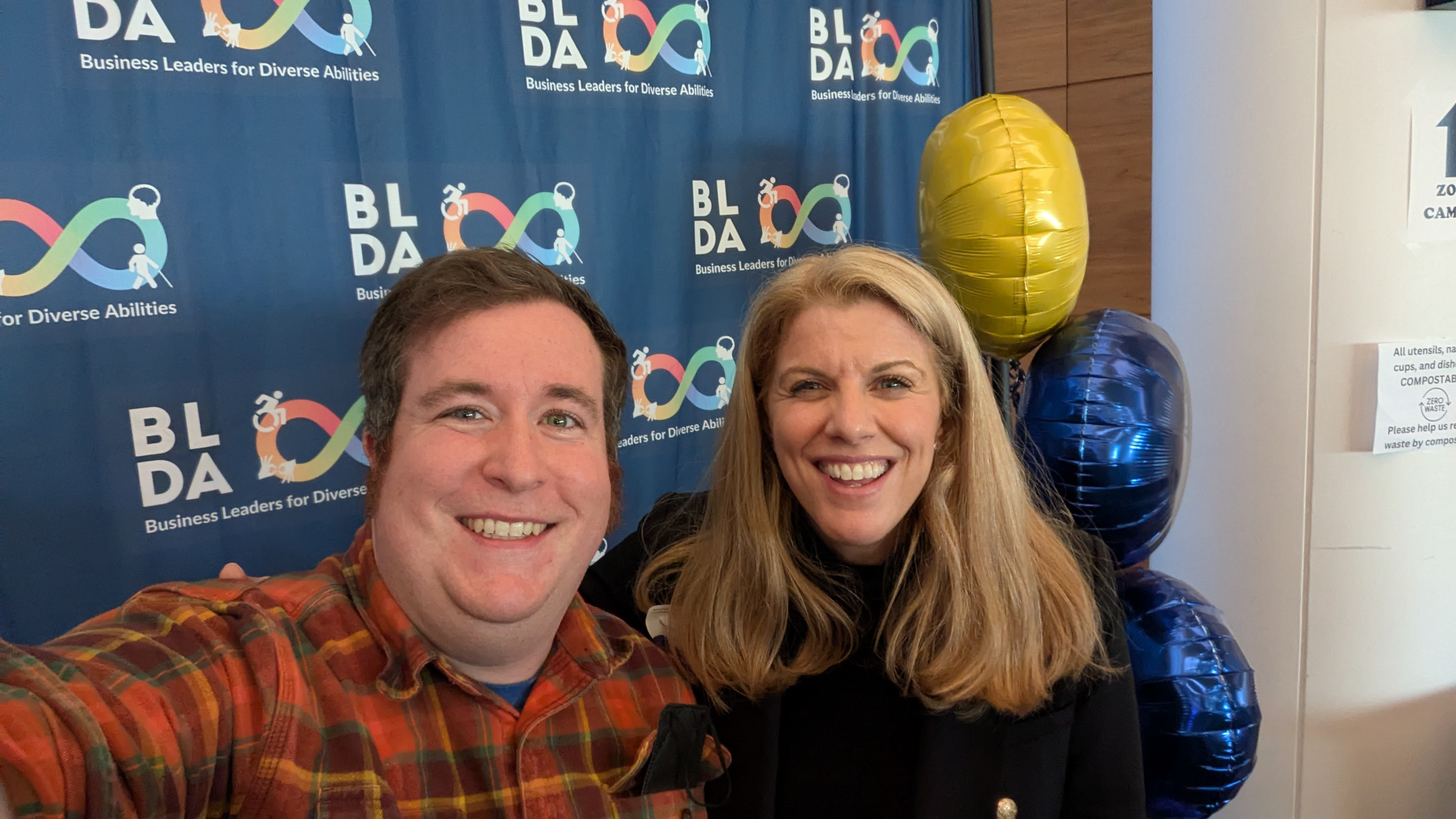 Two smiling individuals, a man and a woman, taking a selfie at an event. The background has a blue banner with the logo and text 'BADA' and 'Business Leaders for Diverse Abilities' repeated multiple times. There are colorful balloons, including a lar