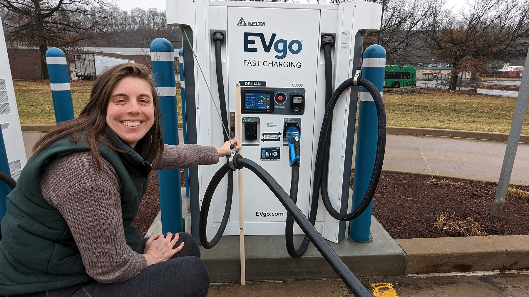A woman smiling and pointing at an electric vehicle charging station outdoors with trees and a street in the background.