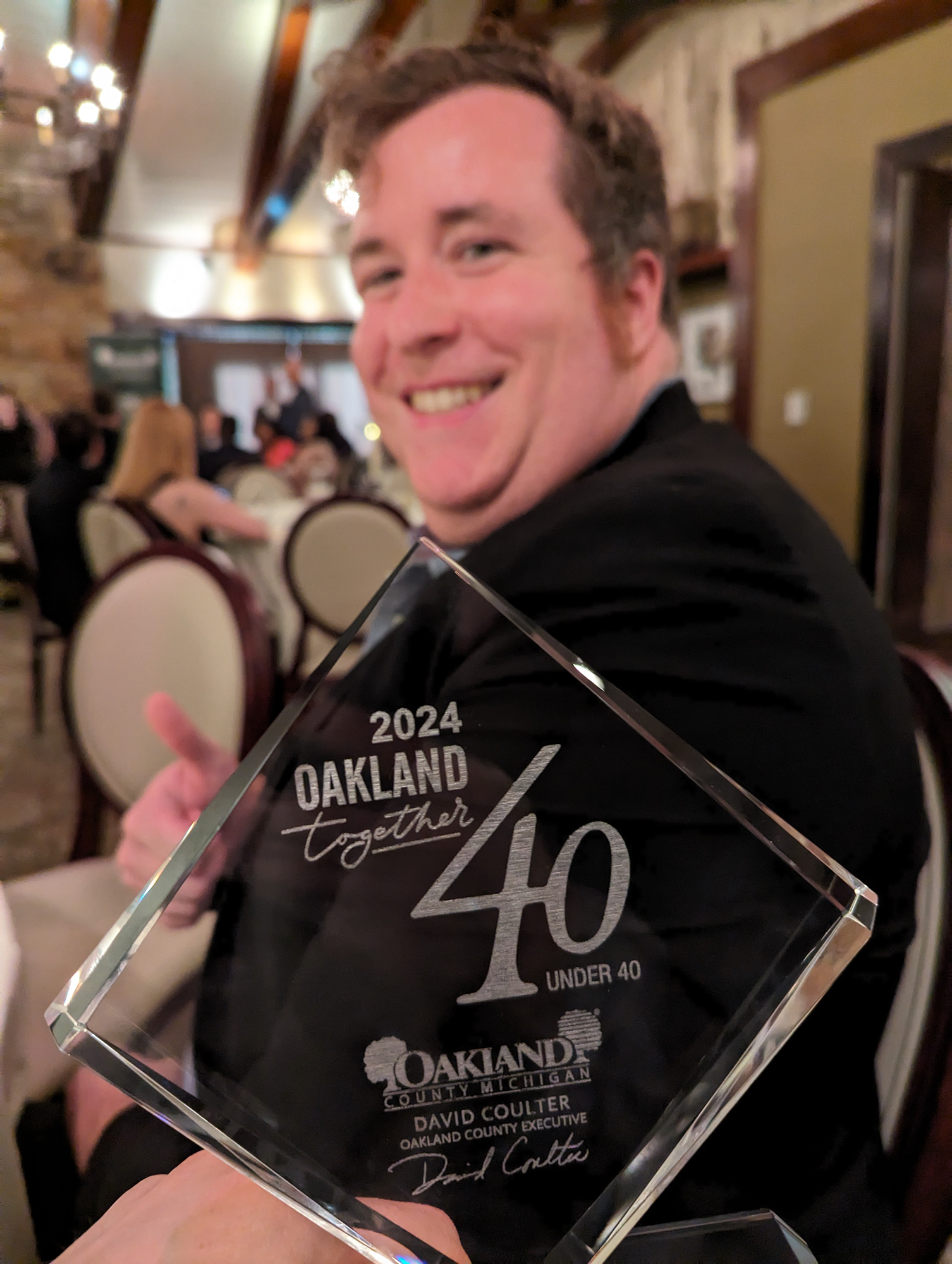 A smiling man at an awards ceremony holding a glass trophy inscribed with the year 2024 and the words Oakland County Michigan. The background shows a restaurant or banquet hall setting with blurred people and warm lighting.