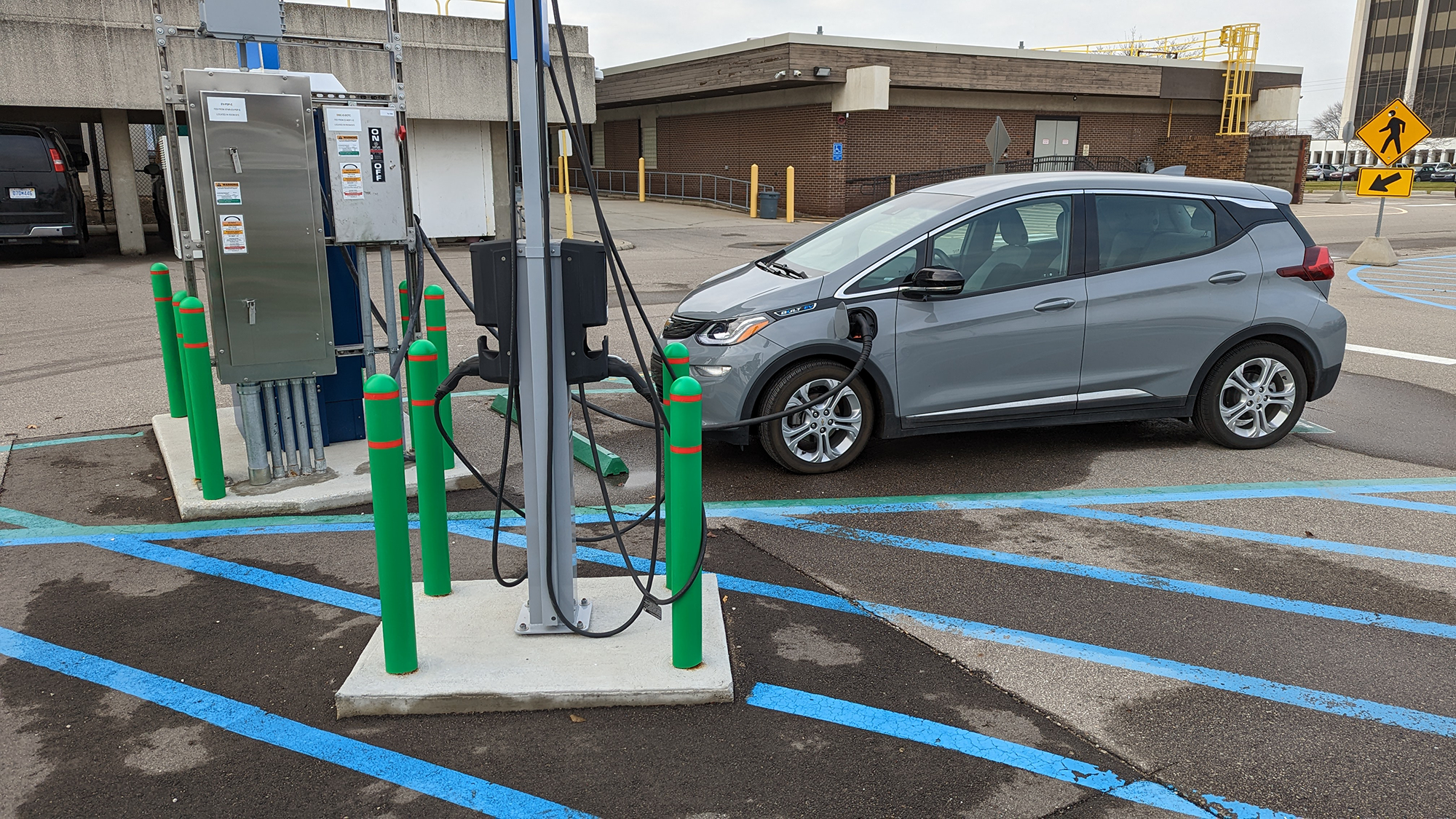 An electric car plugged into a charging station in a parking lot. The car is a gray hatchback, and the charging station has multiple cables and safety poles. The parking lot has blue lines and a handicap space is nearby. There are buildings and signs