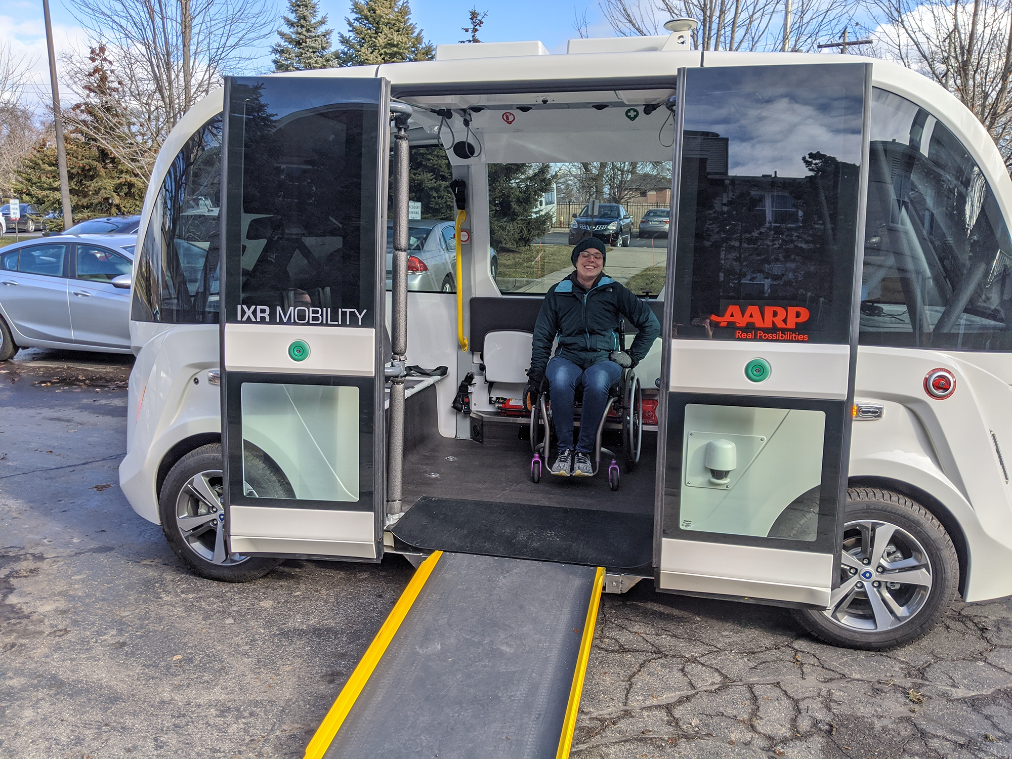 A smiling woman in a wheelchair is going for a ride on an autonomous vehicle, whose doors are open, showing her smiling and still in her chair within the vehicle.