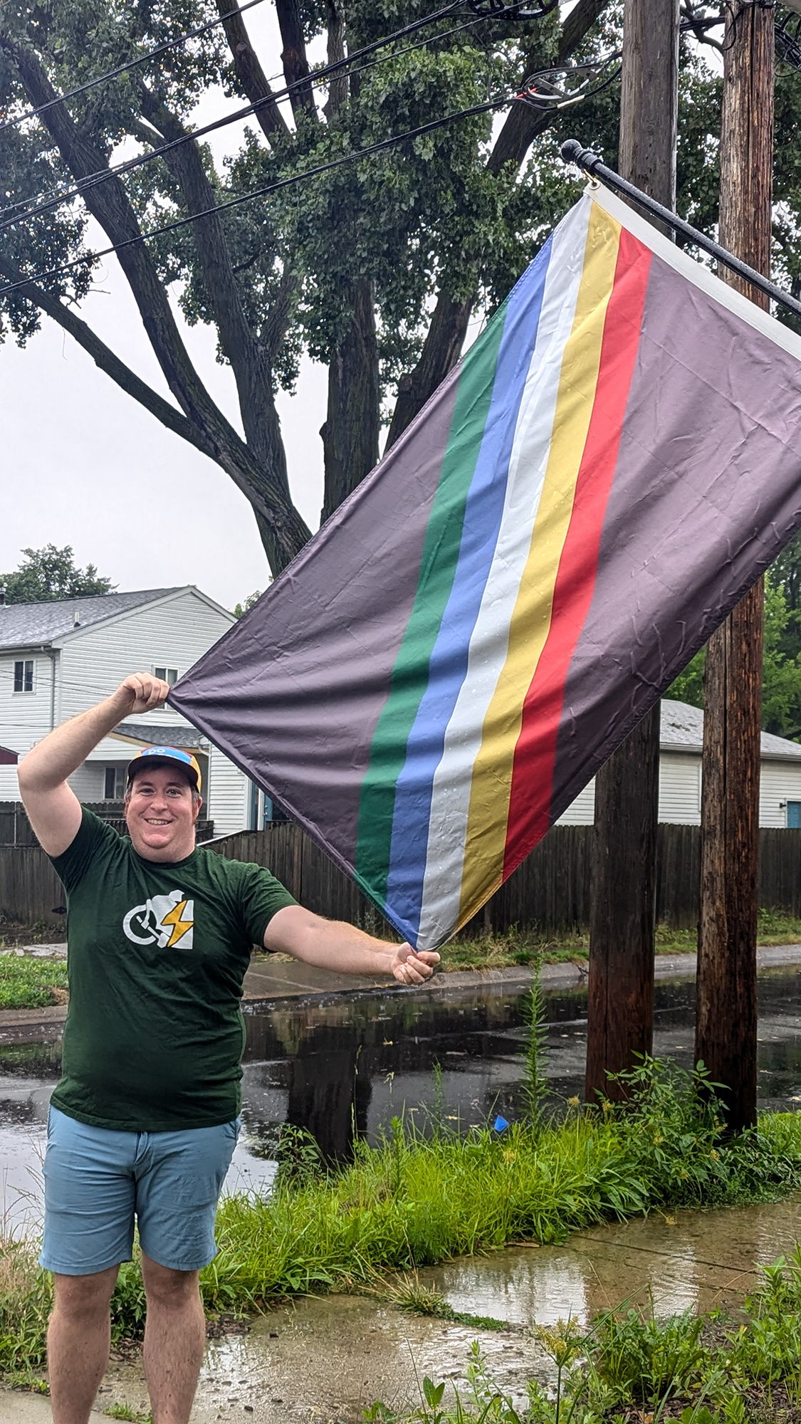 A man smiling and holding a rainbow pride flag outdoors on a rainy day, with a flooded street, trees, and houses in the background.
