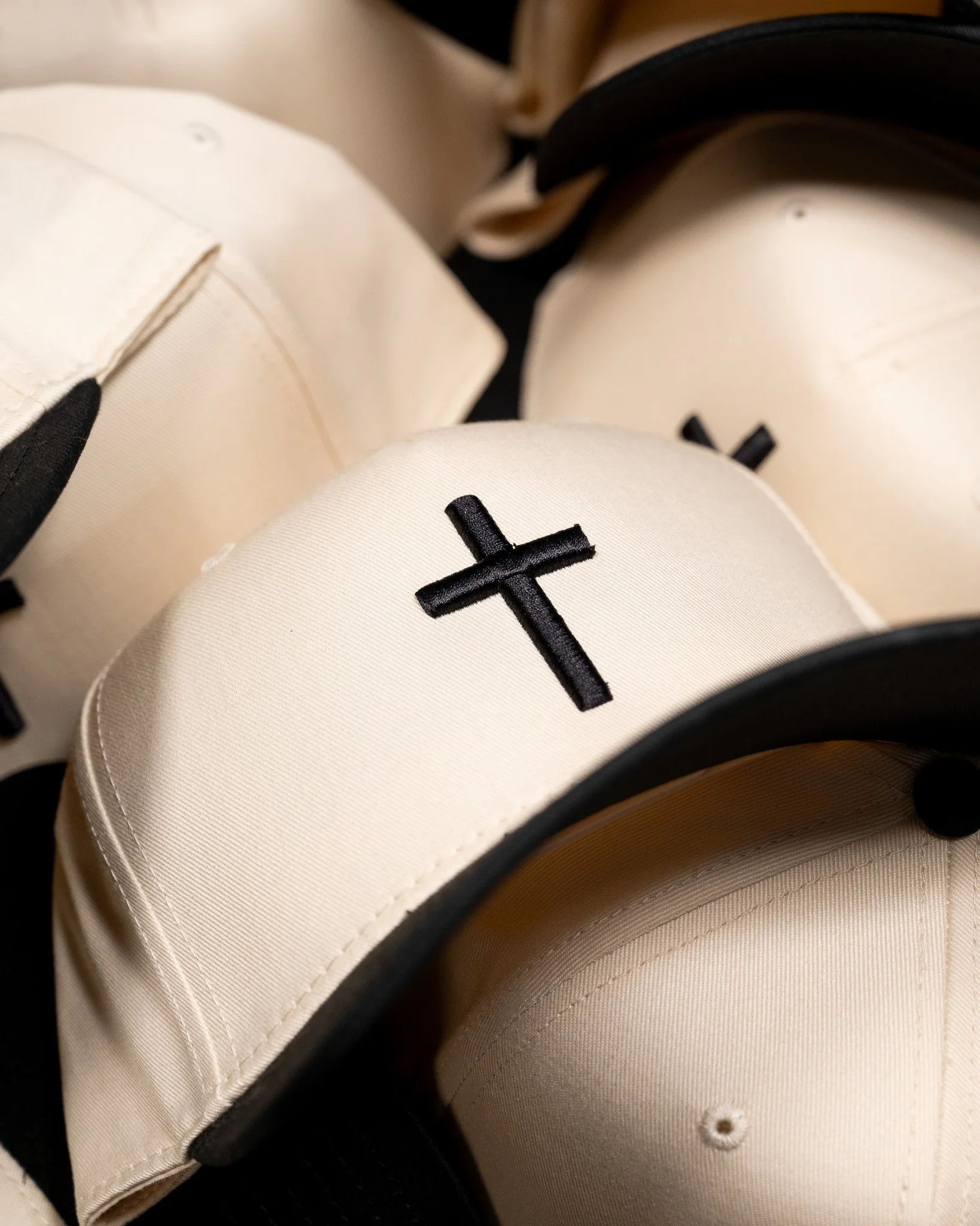 Close-up of a stack of beige baseball caps with black embroidered crosses on the front, some with black trim around the brim. Product photography, flat lay images.