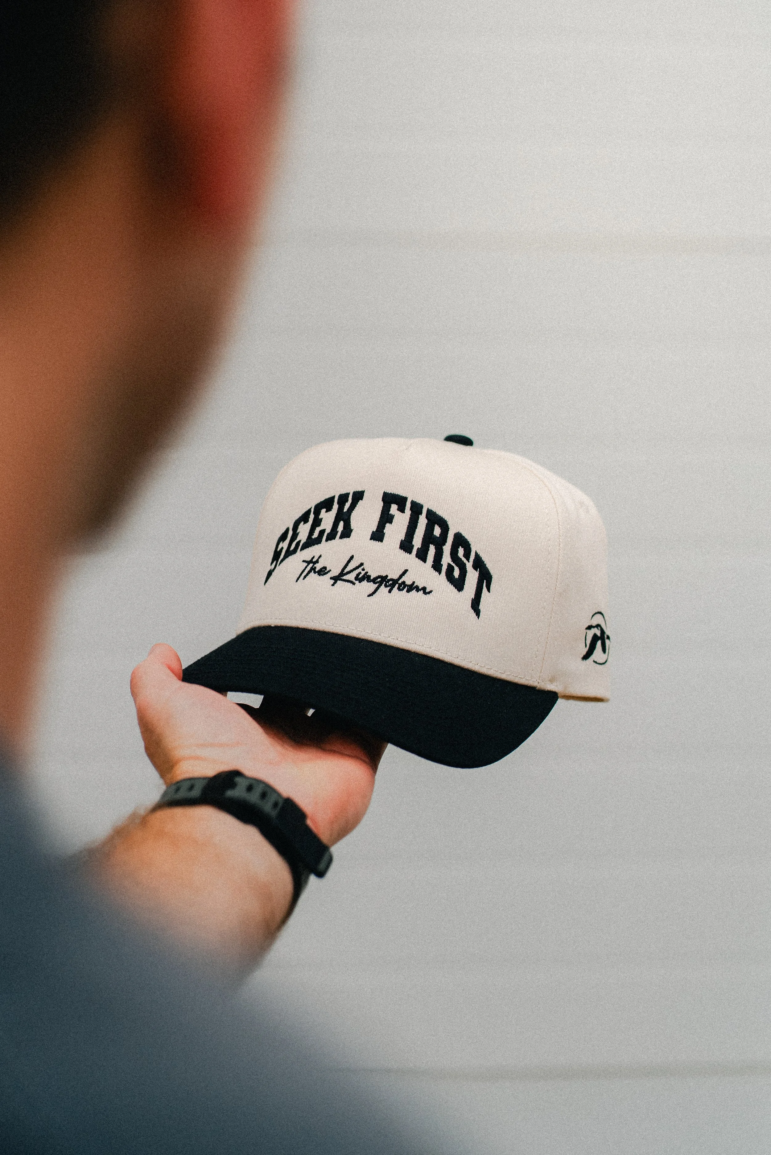 A person holding up a beige and black baseball cap that has the words "GEEK FIRST" and "The Kingdom" embroidered on the front.