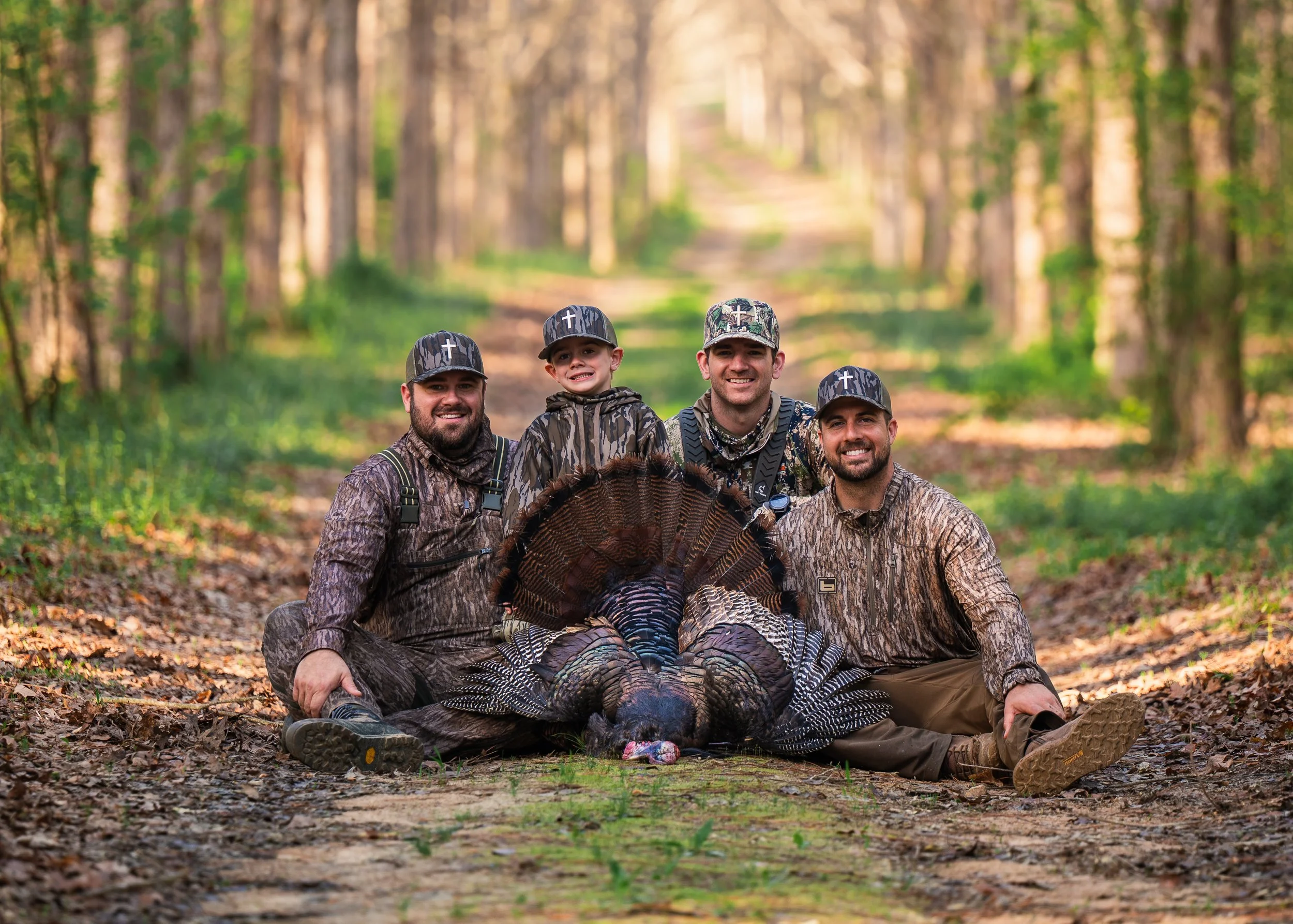 Four men and one boy dressed in camouflage and hunting gear in a forest, sitting on the ground around a hunted turkey. Outdoor lifestyle hunting content creation.