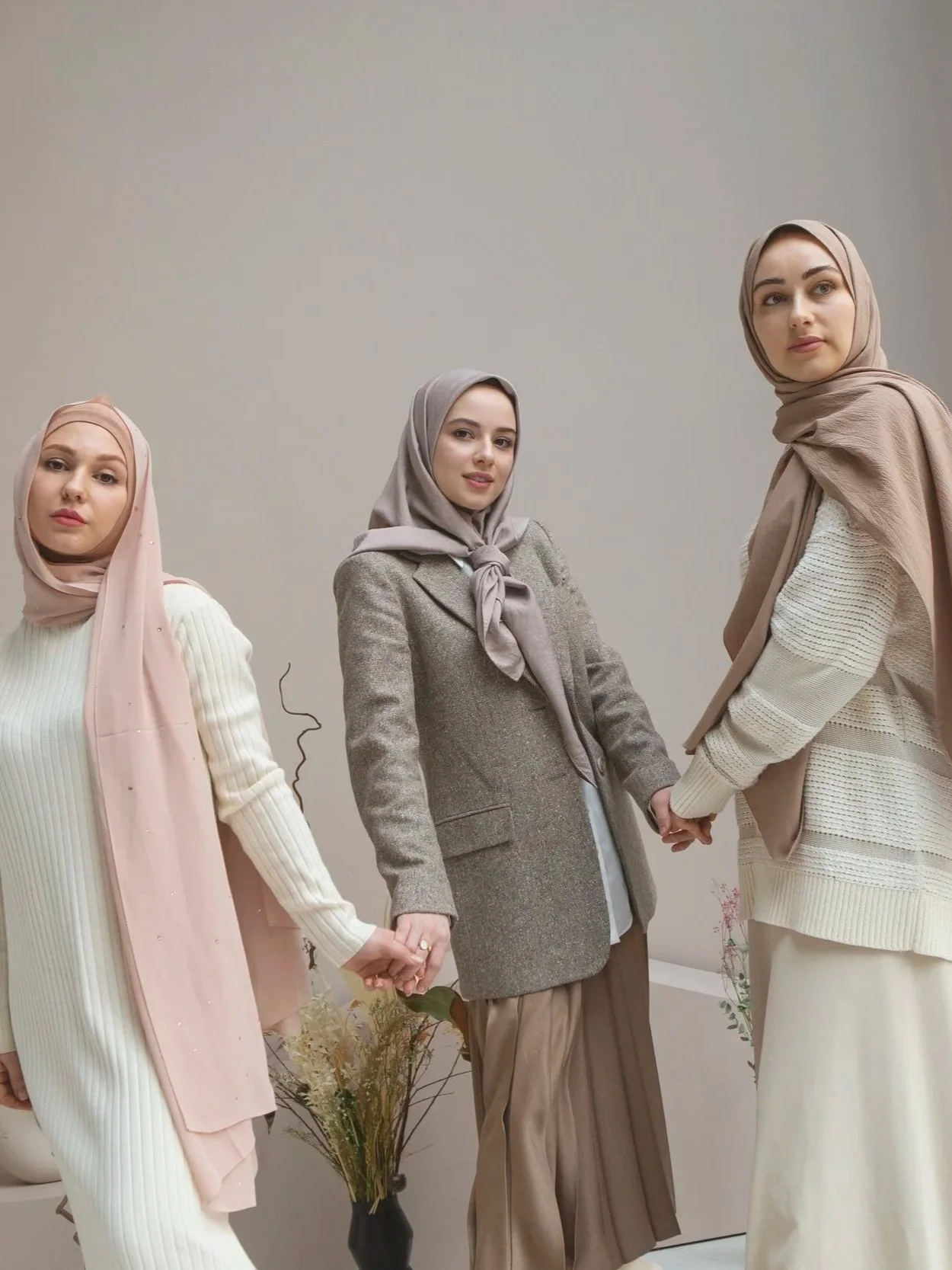 Amnad International. Three women wearing hijabs and modest clothing holding hands in a line, standing indoors with a plain wall background and a flower vase at the bottom left.
