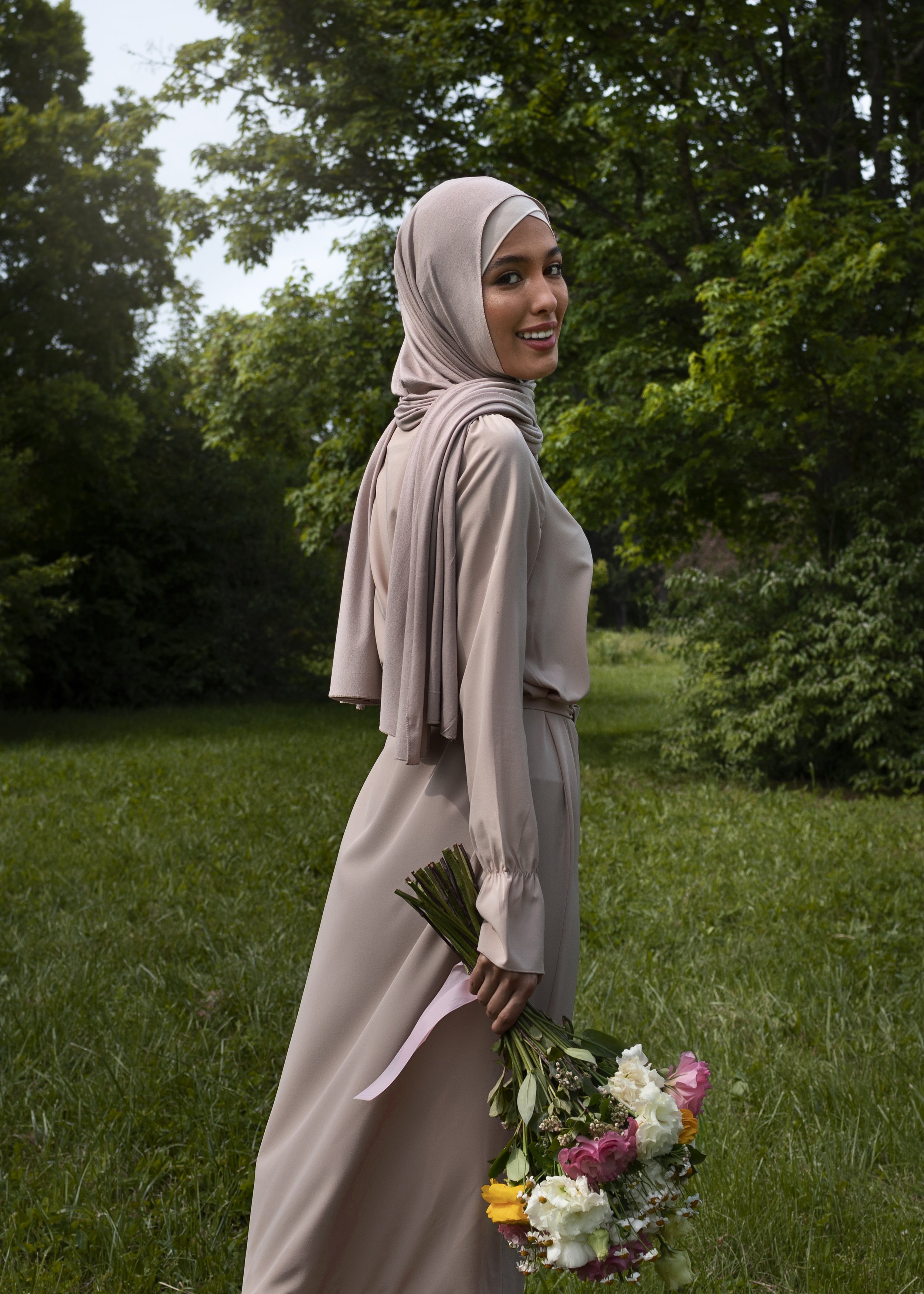A woman in a beige hijab and matching dress standing outdoors in a grassy area with trees in the background, holding a bouquet of colorful flowers.
Amnad International