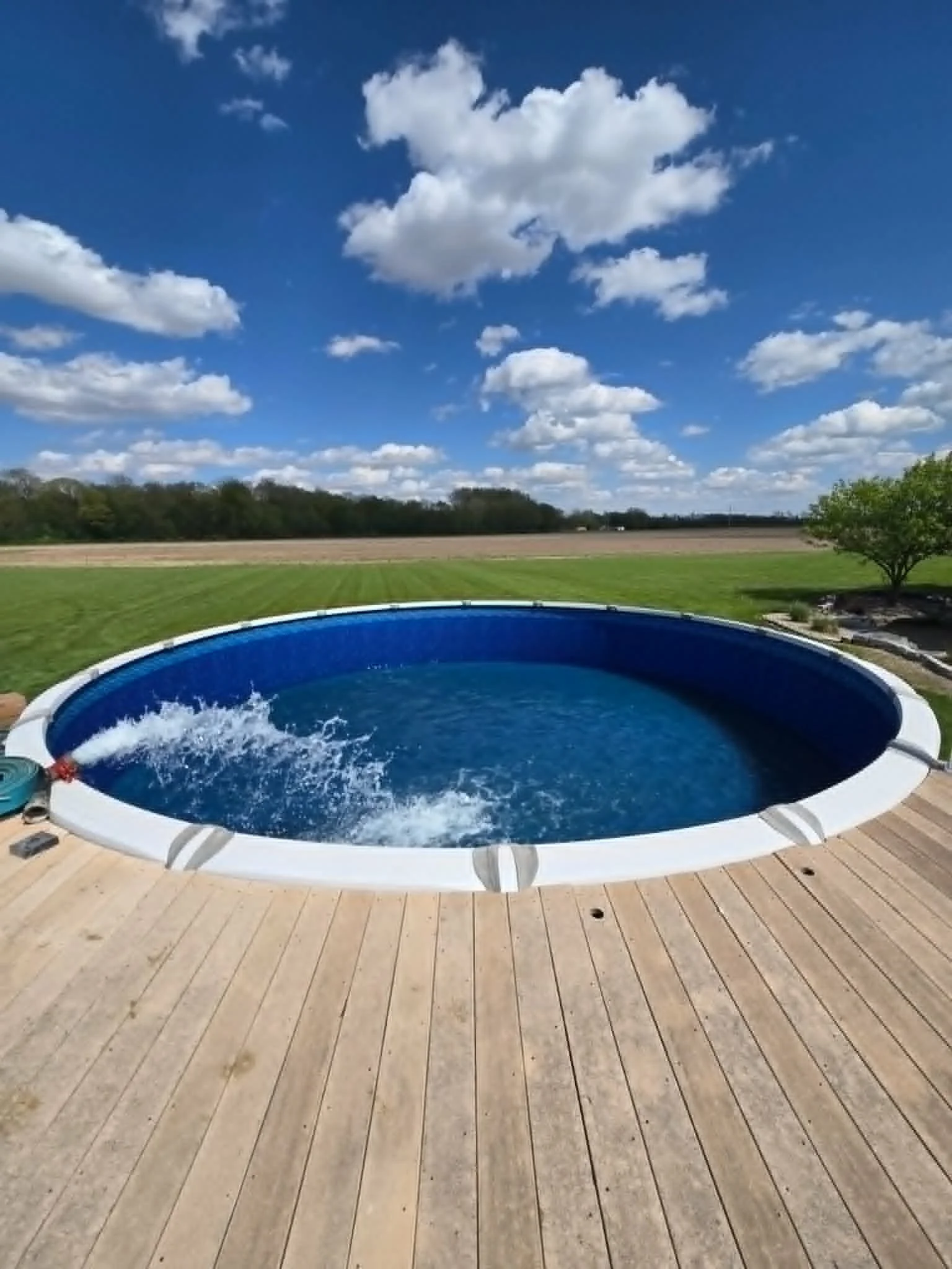 A round outdoor swimming pool filled with water, situated on a wooden deck, overlooking a grassy lawn, trees, and a blue sky with scattered white clouds.